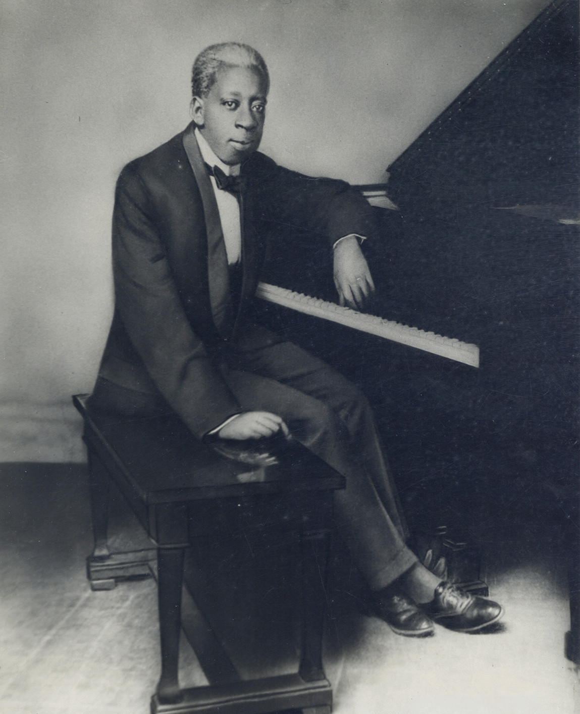 Black and white portrait of a man in a tuxedo seated at a piano bench, one arm resting on the piano keys. He faces the camera with a composed expression. Artifact from the exhibition "The District: The Music and Musicians of Storyville."