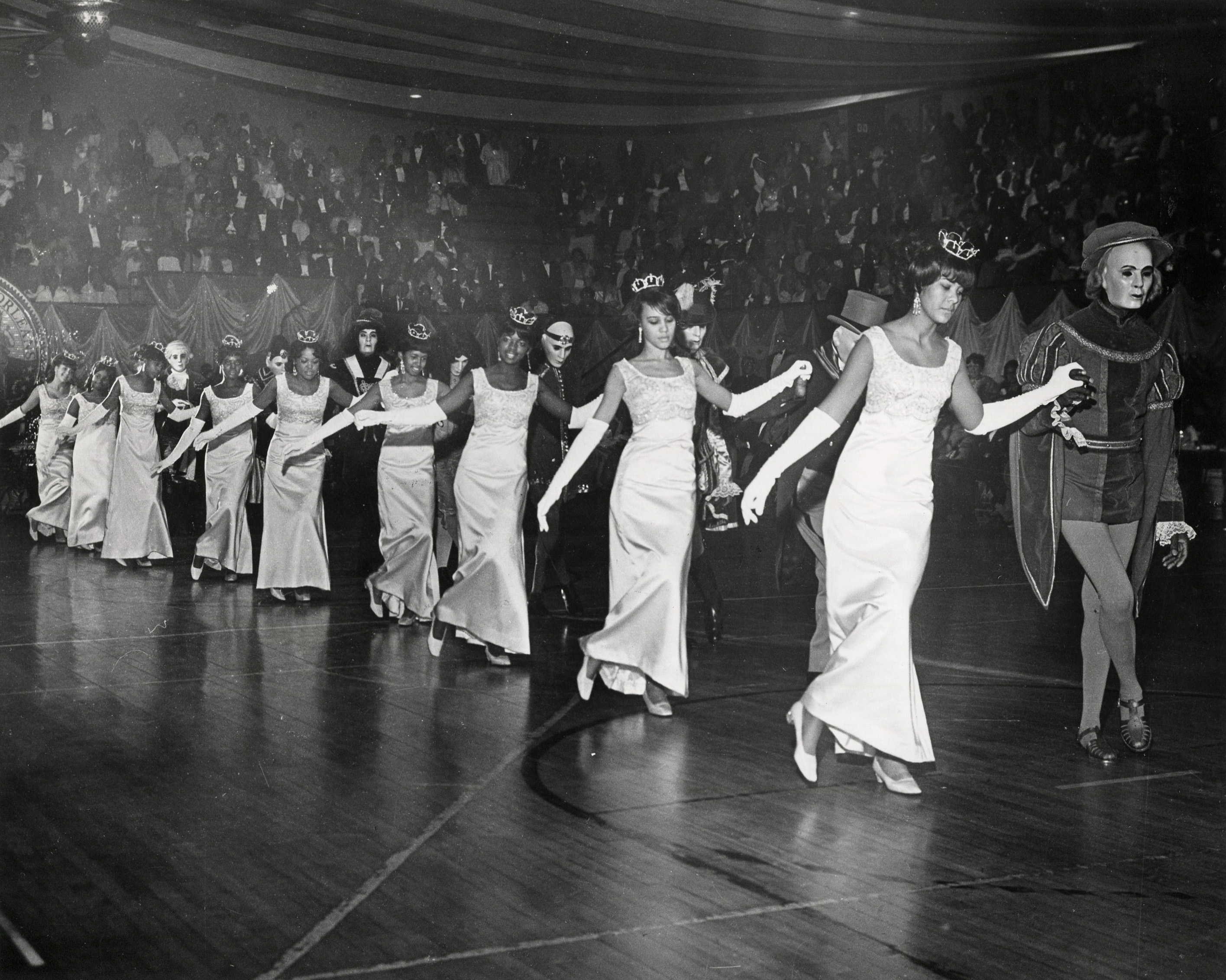 Black-and-white photograph of a line of women in gowns and tiaras, wearing long white gloves, dancing in formation with partners dressed in costumes. The audience is visible in the background watching the ball performance. Original Illinois Club debutantes and members dancing the Chicago Glide, 1968. Photograph by Porter’s Studio.