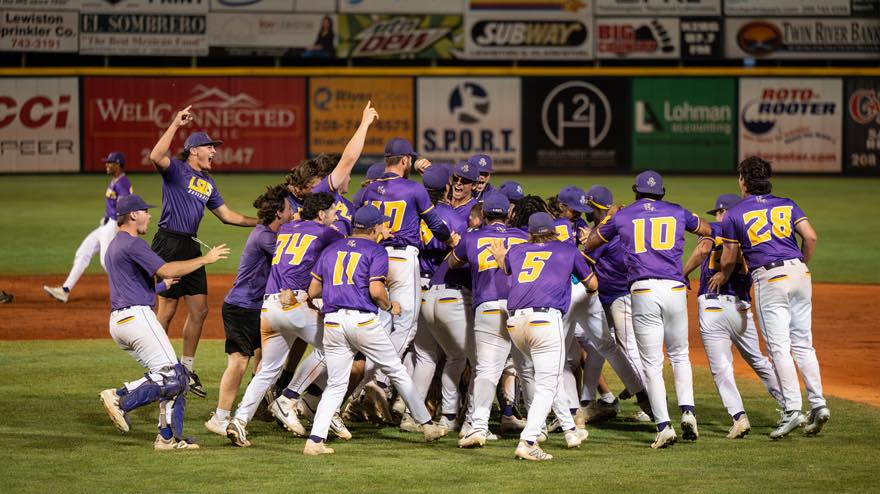 Members of the LSU Shreveport Pilots baseball team wearing purple and gold uniforms celebrate together on the field, cheering and embracing after a victory under stadium lights. Advertising signs line the outfield wall in the background.