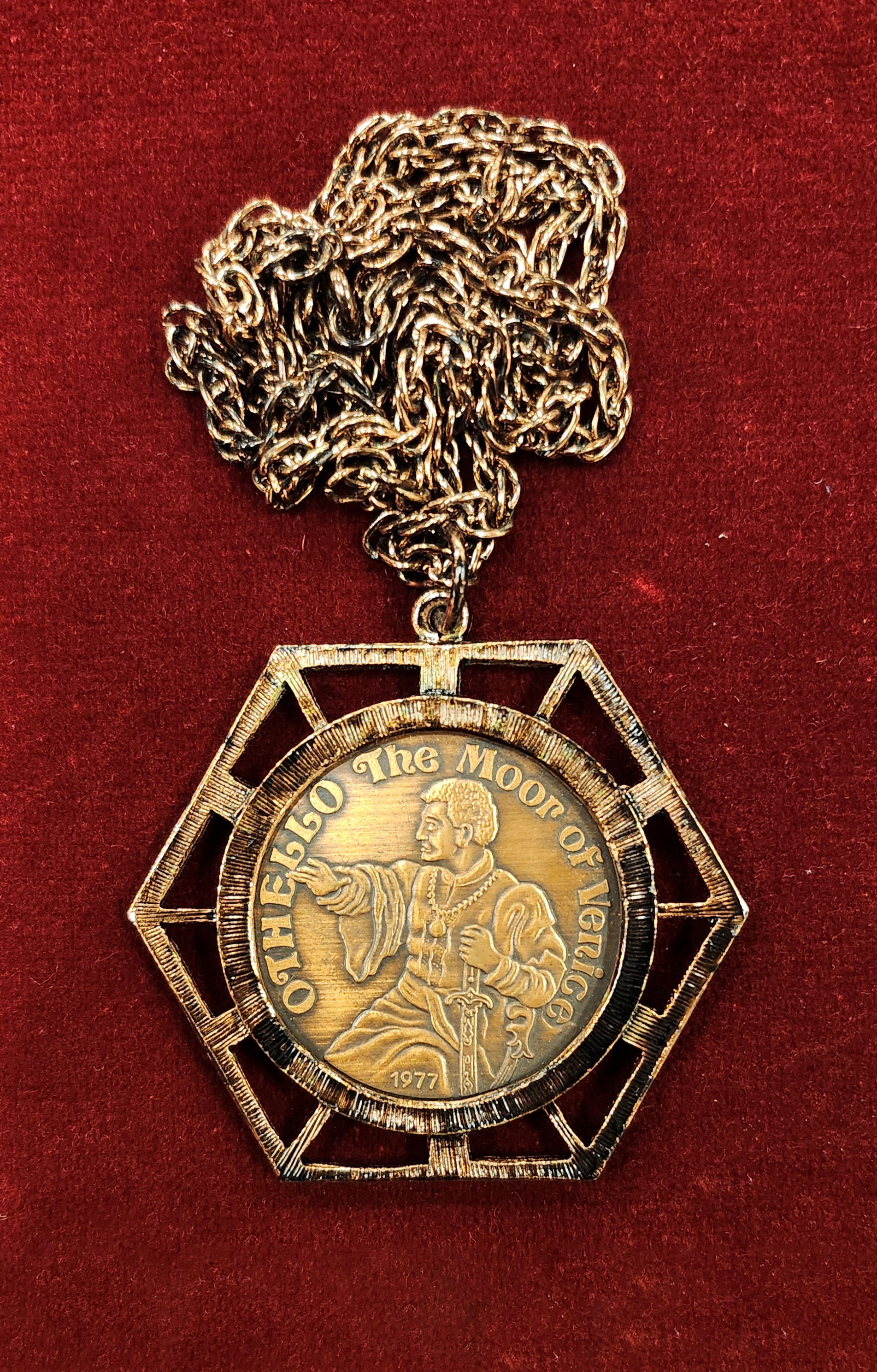 Close-up of a gold medallion on a chain against a red background. The medallion reads “Othello The Moor of Venice, 1977” and shows a raised image of a man in Renaissance clothing pointing outward. Member’s medallion, Original Illinois Club, 1977.