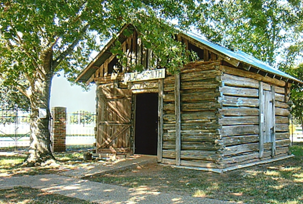 Photograph of a small rustic wooden blacksmith shop with a tin roof and a sign reading “Blacksmith” above the doorway. The log structure sits under the shade of large trees, with a brick fence and grassy yard in the background.