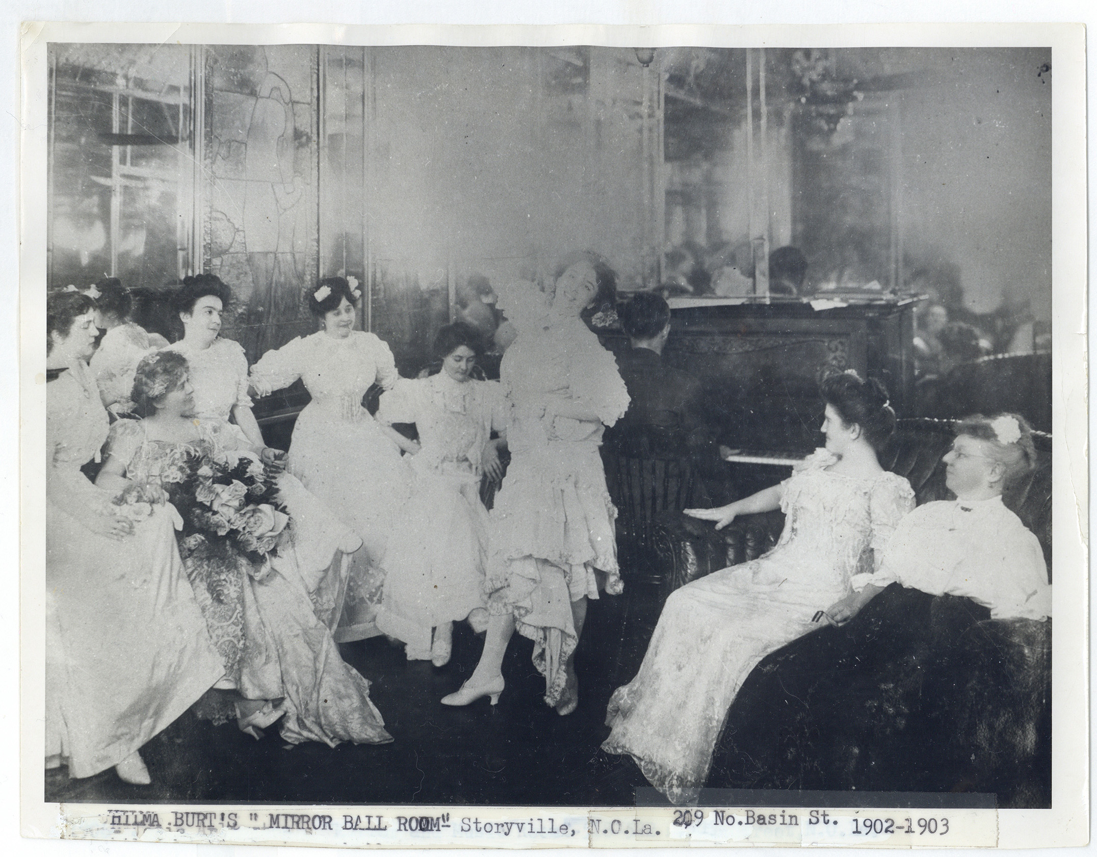 Black-and-white photograph of women in elegant early 1900s gowns gathered in a parlor known as “Emma Burt’s Mirror Ball Room” in Storyville, New Orleans, around 1902–1903. One woman dances while others sit and watch, surrounded by ornate mirrors, stained glass, and a piano in the background. The scene captures the social atmosphere of New Orleans’s Storyville era.