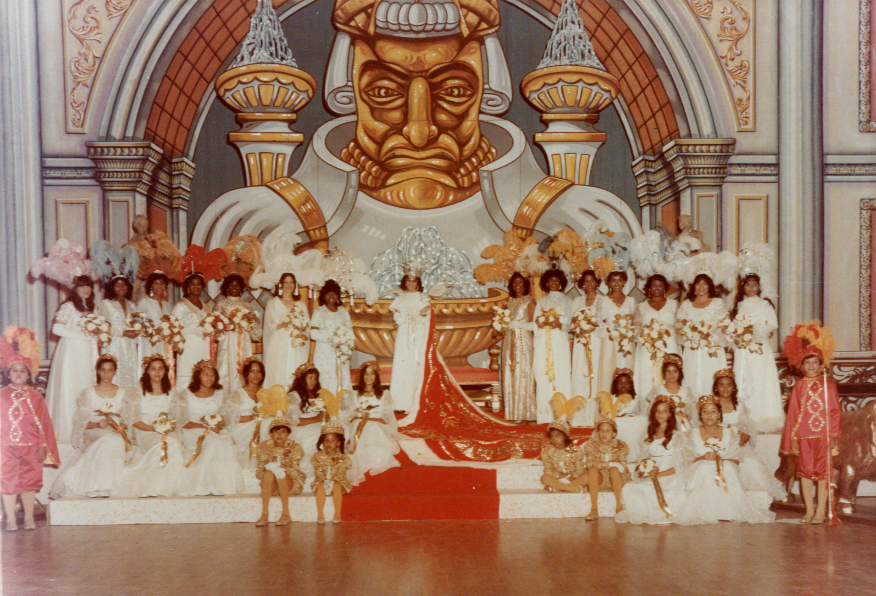 Color photograph of a Mardi Gras court. A queen in a white gown with a long red train stands on a platform surrounded by maids of honor in white dresses with feathered headpieces, holding bouquets. Pages in gold costumes and young attendants sit at the front of the stage. A large painted backdrop of a stylized regal figure dominates the background. Court of the Young Men Illinois Club, 1974.