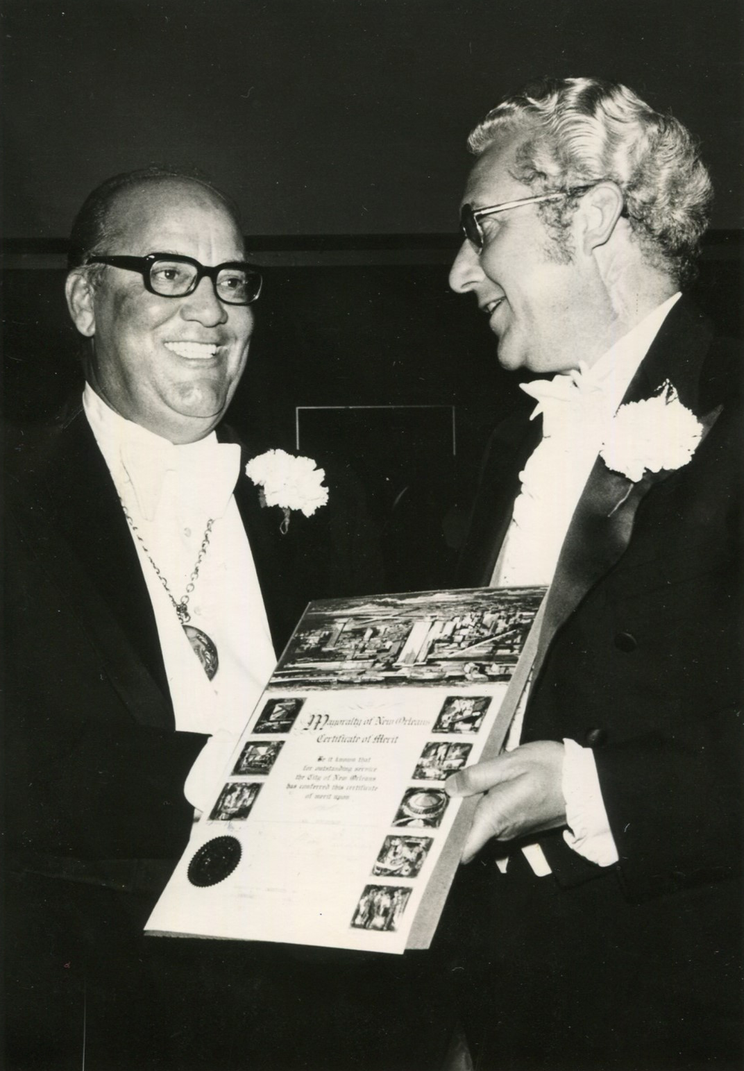 Black-and-white photograph of two men in tuxedos smiling and talking. One man is holding a certificate of merit from the City of New Orleans decorated with small illustrations. Carl Duvigneaud, Ball Chairman of the Original Illinois Club, presented with a certificate of recognition by New Orleans Mayor Moon Landrieu, 1970s.