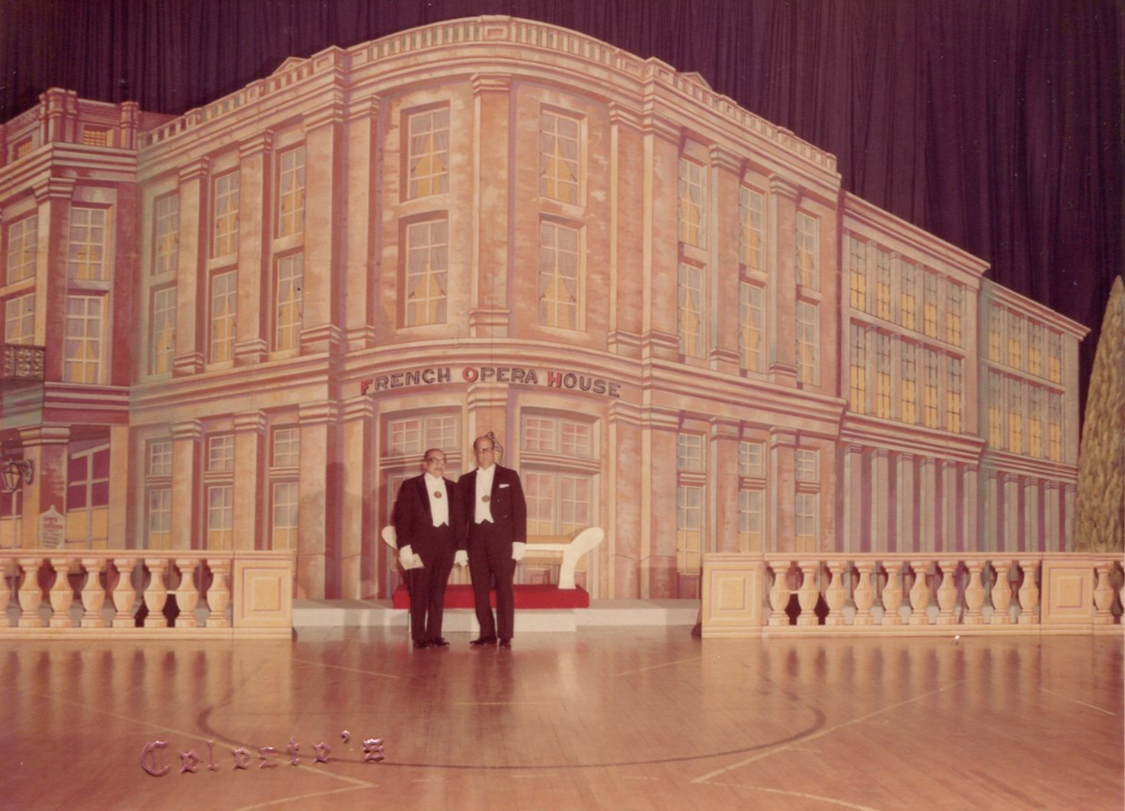 Color photograph of two men in tuxedos standing on a stage in front of a large painted backdrop depicting the French Opera House. The set includes a red carpet and white balustrades, giving the appearance of a grand entrance. Marcus Neustadter Jr. and Carl Duvigneaud at the Original Illinois Club ball, 1968. Photograph by Celeste Broadway.