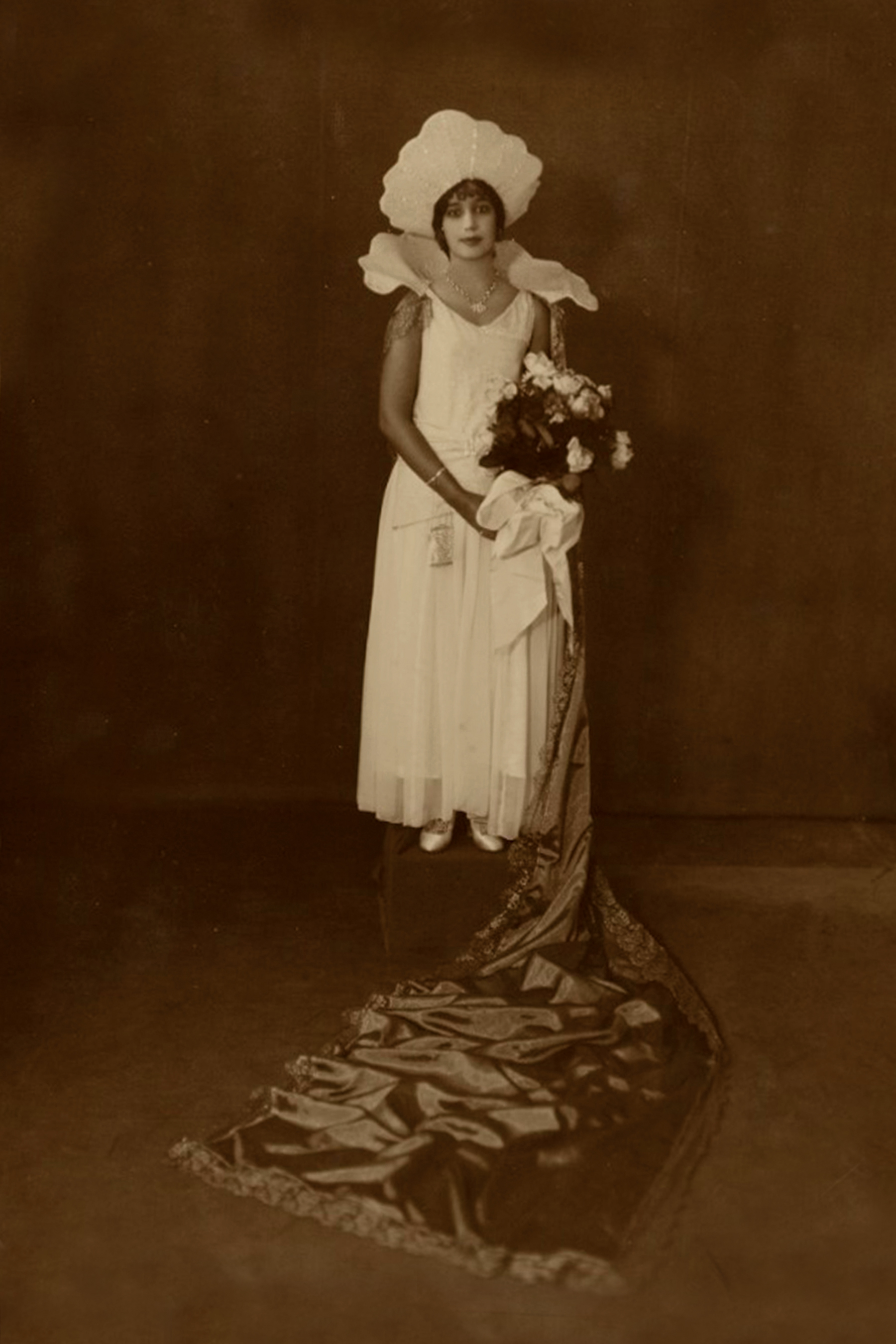 Black-and-white photograph of a line of women in gowns and tiaras, wearing long white gloves, dancing in formation with partners dressed in costumes. The audience is visible in the background watching the ball performance. Verlie Bright, Queen of the Young Men Illinois Club, 1930. Photograph by Villard Paddio.