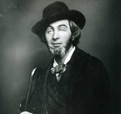 Black-and-white portrait of a bearded man wearing a dark coat, wide-brimmed hat, and bow tie, seated and holding a cane. The image shows poet Walt Whitman in his younger years, gazing calmly at the camera.