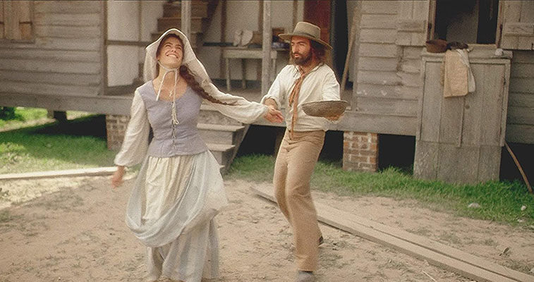 A woman in 19th-century Cajun clothing smiles and spins outdoors while a bearded man in period attire reaches toward her with a wooden bowl in his hands. They stand in front of a weathered wooden house raised on brick piers.