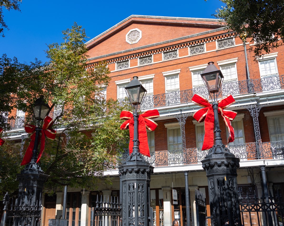 Holiday decorations in Jackson Square: Three black iron lampposts topped with bright red bows stand in front of the historic Pontalba Building, with its red brick façade, lacy wrought-iron balconies, and a clear blue sky overhead.