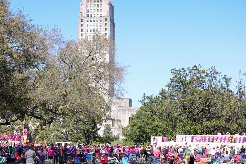 Crowds gathered outdoors during the Spanish Town Parade in Baton Rouge, with spectators wearing colorful clothing and costumes seated in folding chairs and standing along the route, trees lining the scene, and the Louisiana State Capitol tower rising prominently in the background under a clear blue sky.