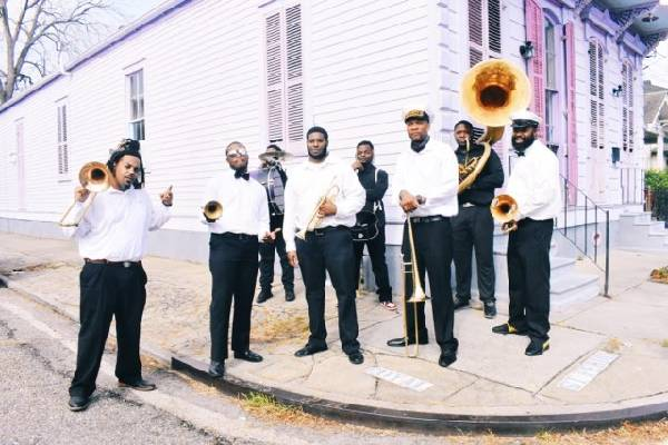 Sporty’s Brass Band poses outdoors on a New Orleans street corner, wearing black-and-white attire and holding brass instruments including trumpets, trombones, and a sousaphone, with colorful historic buildings in the background.