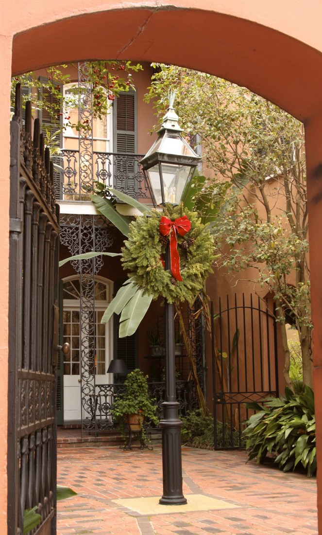 Photograph of a traditional New Orleans Creole courtyard framed by a stucco archway. At the center stands a black iron gas-style lamppost decorated with a green holiday wreath and red bow. Wrought-iron gates, brick paving, potted plants, and a two-story building with decorative iron balconies and shuttered doors surround the intimate courtyard space.