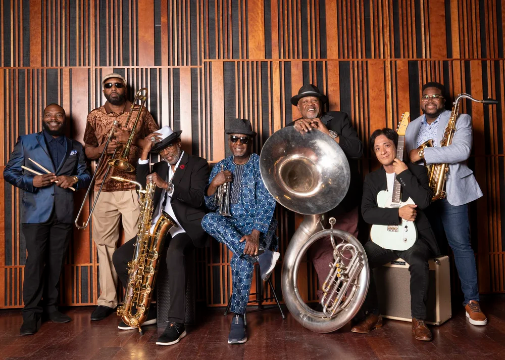 The Dirty Dozen Brass Band poses for a group portrait in a recording studio, with members standing and seated against a wood-paneled wall while holding brass instruments including trombone, saxophones, trumpet, sousaphone, and guitar.