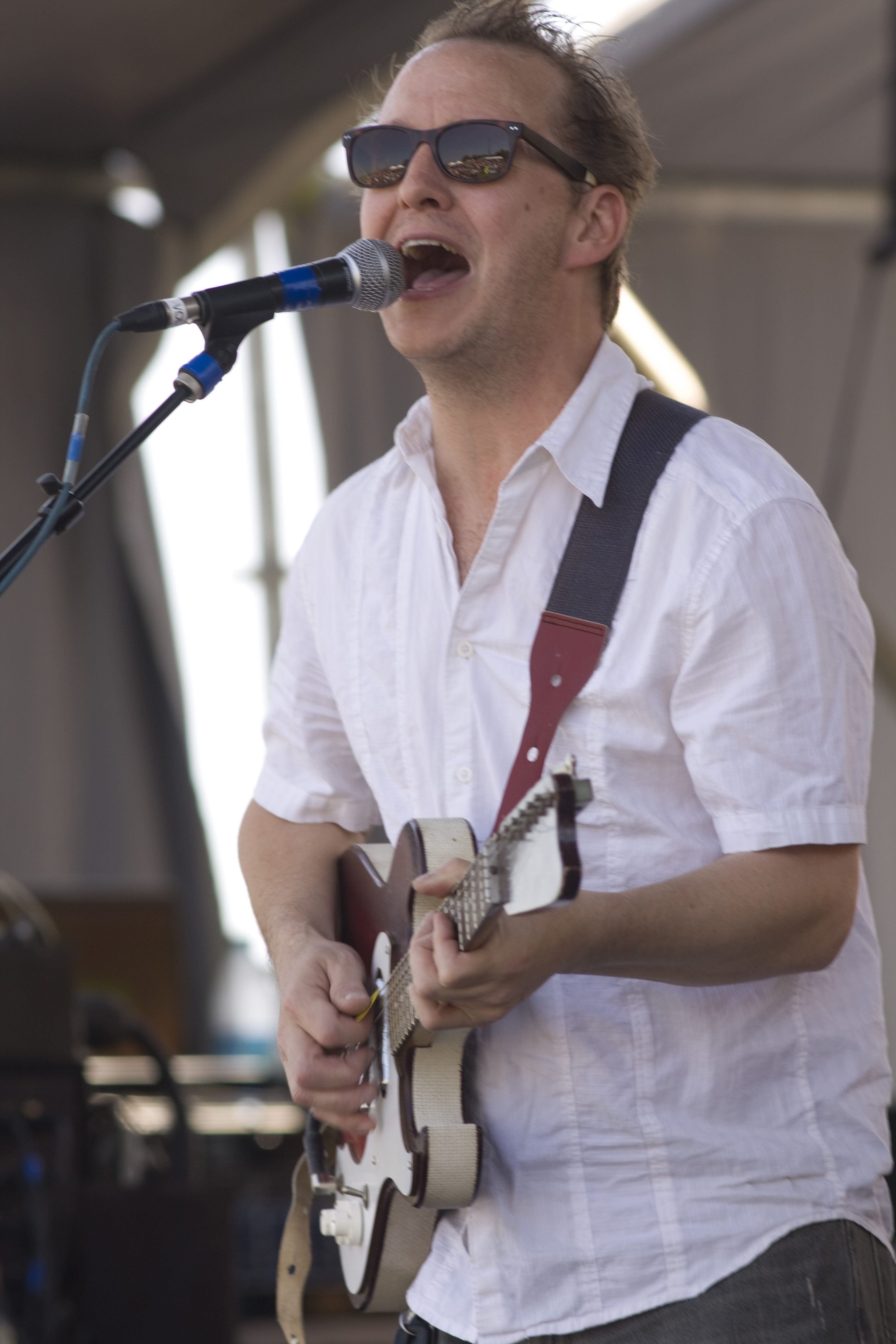 Alex McMurray performs onstage, singing into a microphone while playing an electric guitar. He wears a white short-sleeve shirt and sunglasses, with a guitar strap over his shoulder, during an outdoor live music performance.