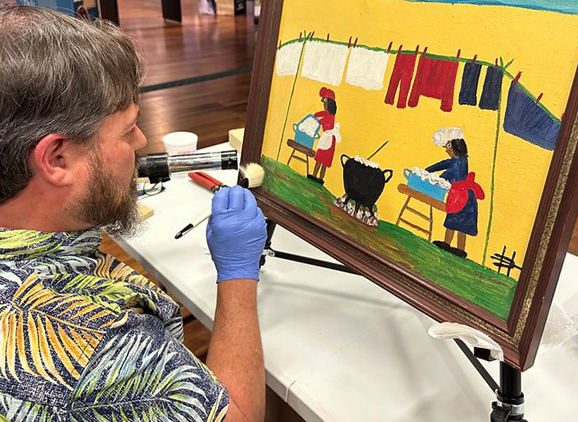 Conservator wearing blue gloves carefully cleans a framed folk-style painting on an easel, using a small brush while seated at a worktable inside a museum gallery.