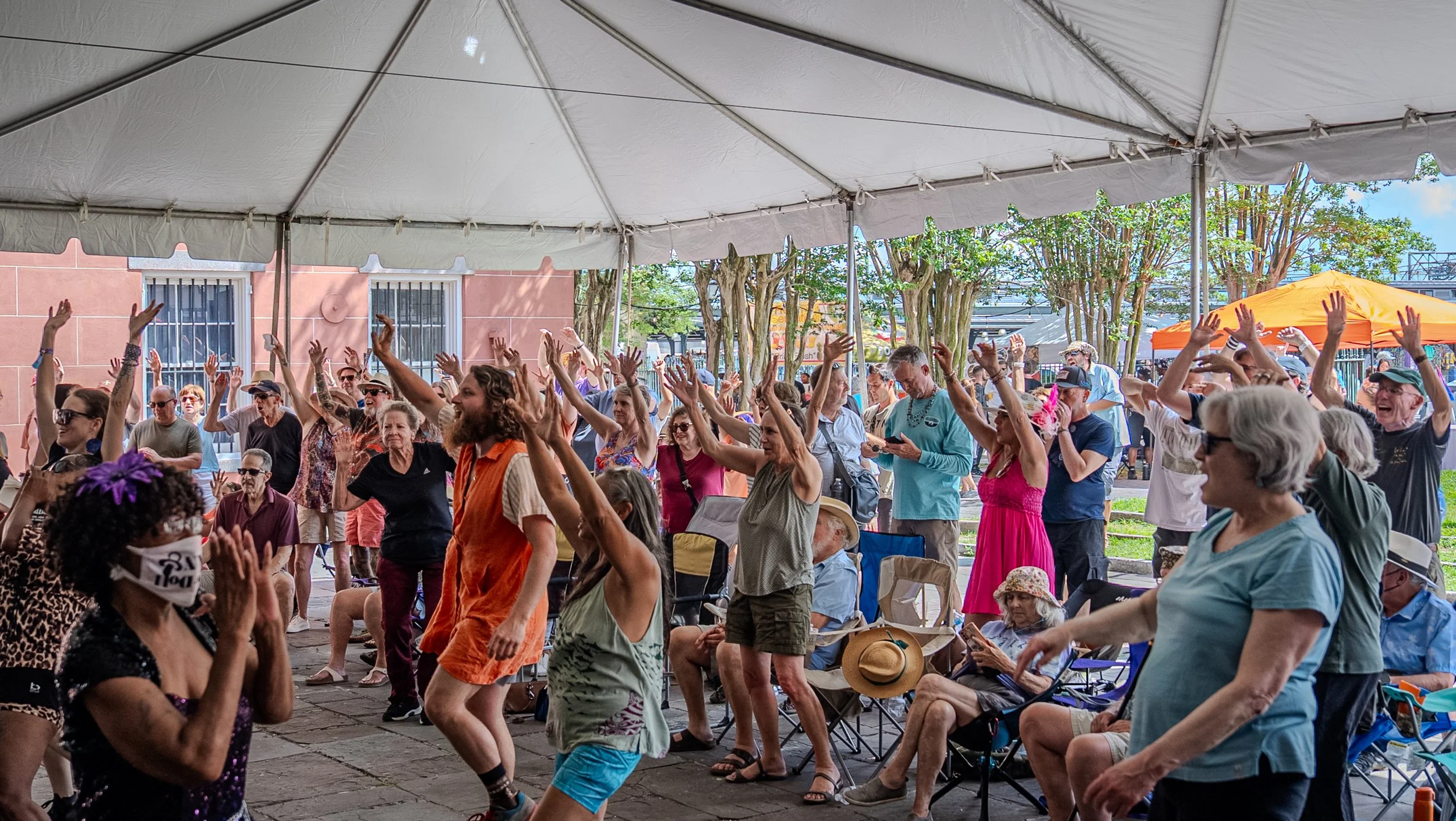 A large crowd of people dances with their hands raised under a white event tent during an outdoor festival. Attendees of various ages fill the space, some standing and some seated in folding chairs, with bright clothing and expressive movement. Behind the tent, trees, vendor tents, and the exterior of a peach-colored building are visible.