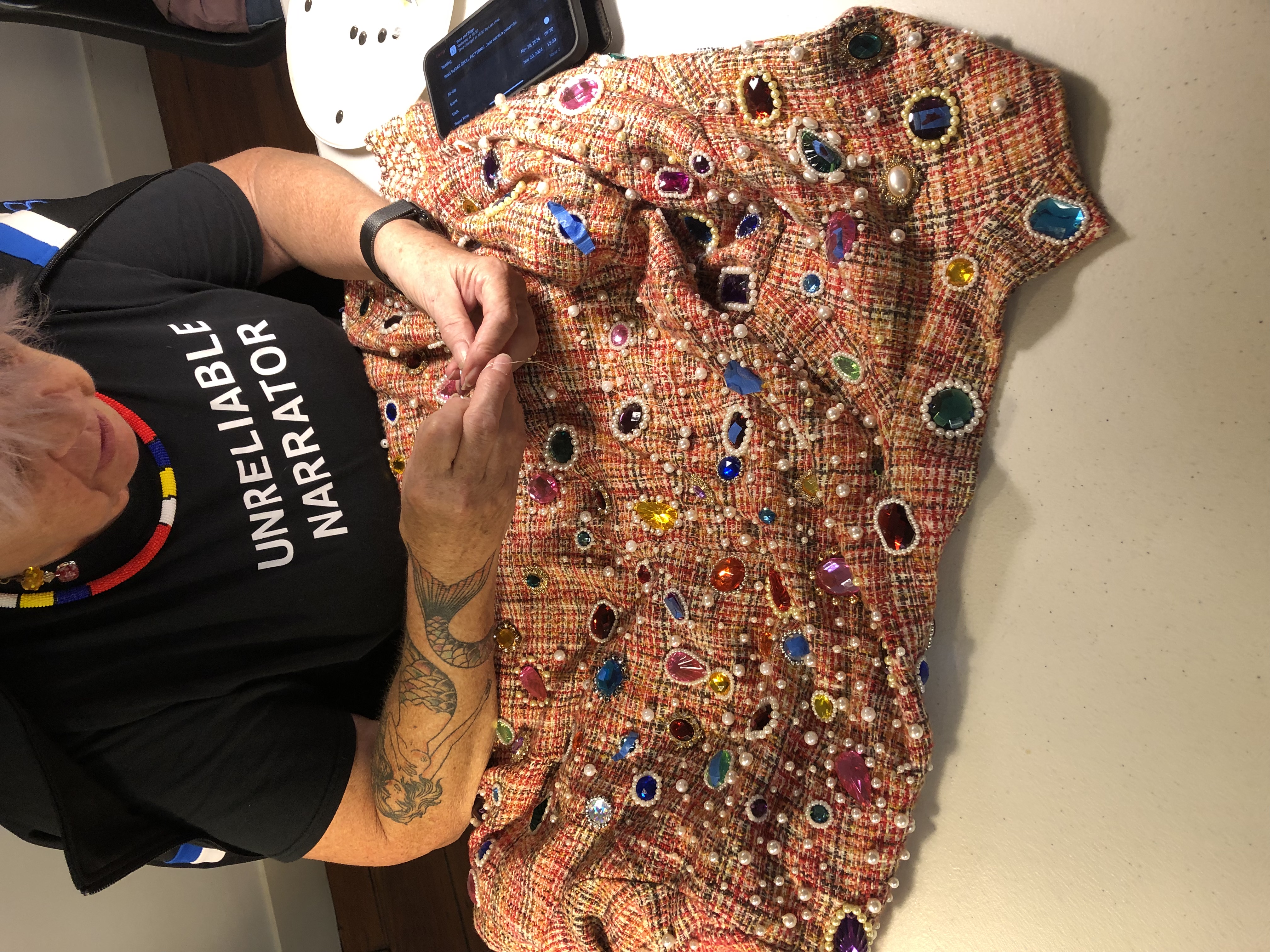 A person wearing a black T-shirt that reads “UNRELIABLE NARRATOR” hand-sews beads and colorful rhinestones onto a textured orange-and-pink fabric. The table is covered with the embellished garment, which is decorated with pearls and multicolored gems. A smartphone sits nearby.