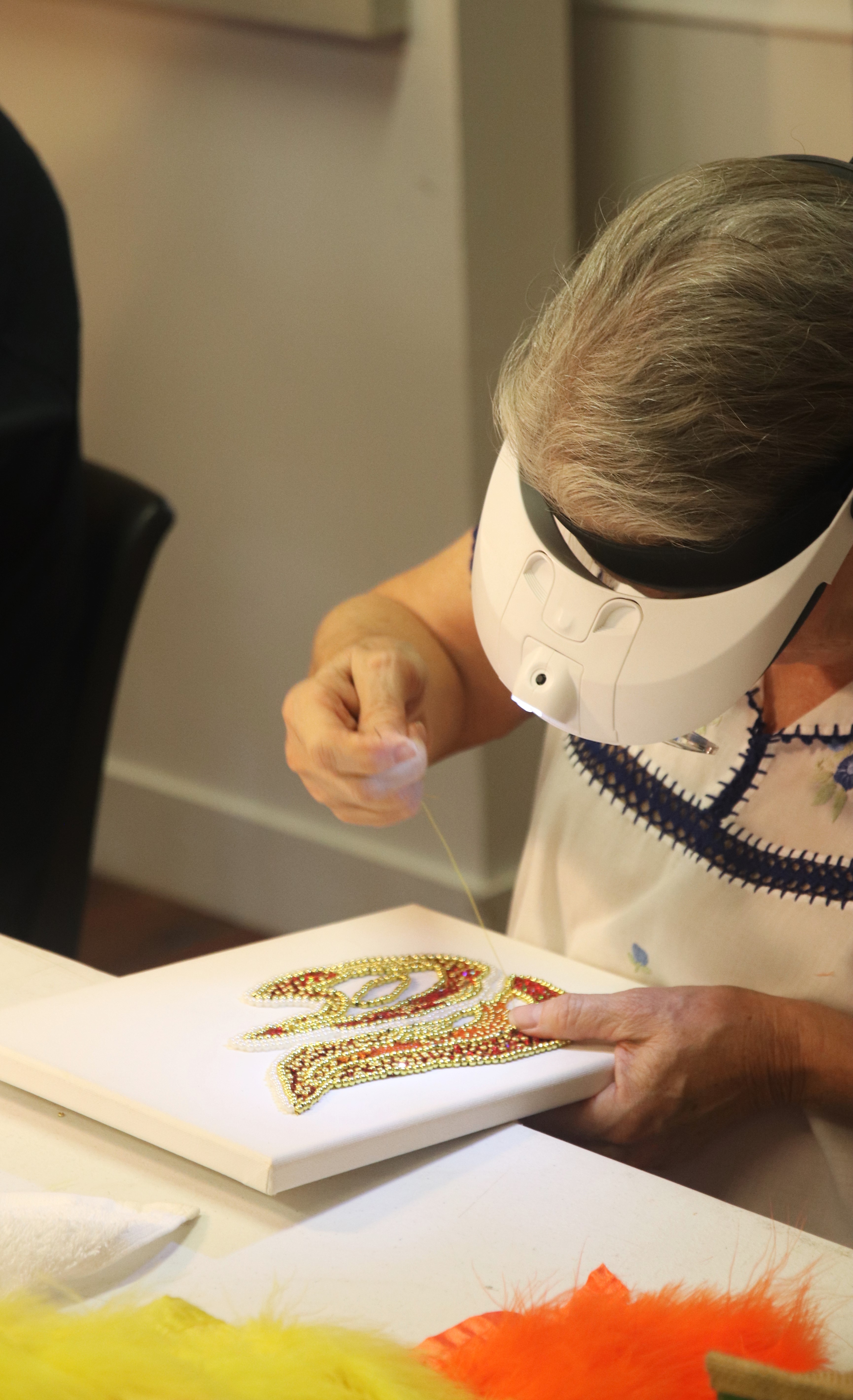 A woman with gray hair, wearing a white embroidered blouse and a magnifying visor, stitches intricate beadwork onto a white canvas. Bright yellow and orange feathers are visible on the table.