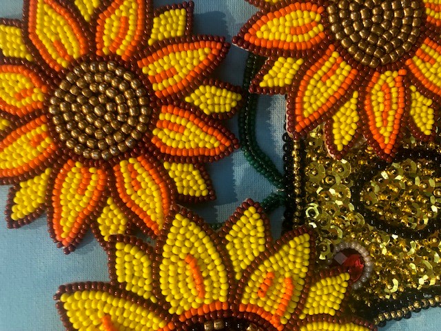 A close-up of beadwork depicting bright orange and yellow sunflowers with brown beaded centers. Sequins and small beads create detailed textures and shading.