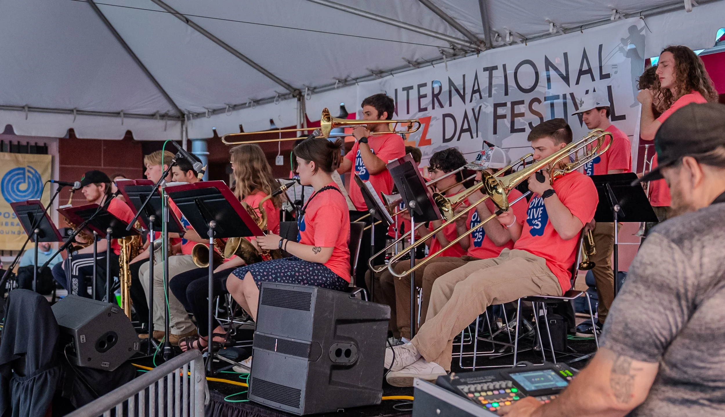 A large student jazz ensemble performs onstage under a white festival tent. Musicians in matching bright coral shirts play trombones, saxophones, and trumpets while reading from music stands. A banner behind them reads “International Jazz Day Festival.” A sound engineer operates a mixing console in the foreground.