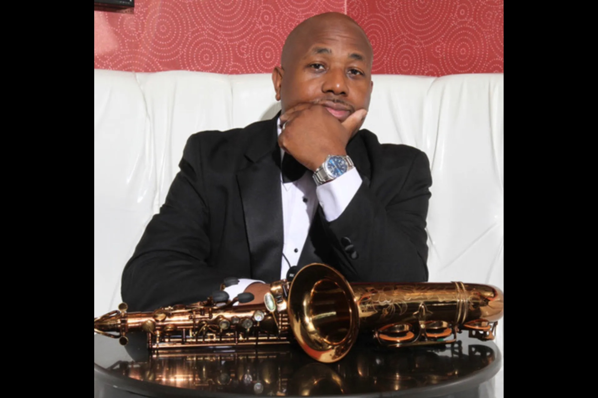 A handsome Black man in a black tuxedo sits on a white leather bench, resting his chin on his hand in a thoughtful pose. A polished brass saxophone lies on the table in front of him, reflecting light. A red patterned wall forms the background.