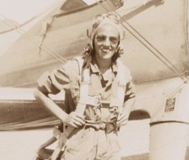 A smiling woman in a flight suit stands in front of an airplane, wearing a parachute harness and aviator gear.