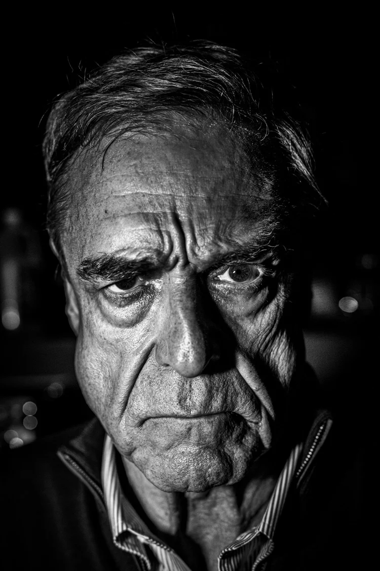 Dramatic black-and-white close-up portrait of John Goodman staring directly at the camera, his face partly shadowed and deeply lined, against a dark background.