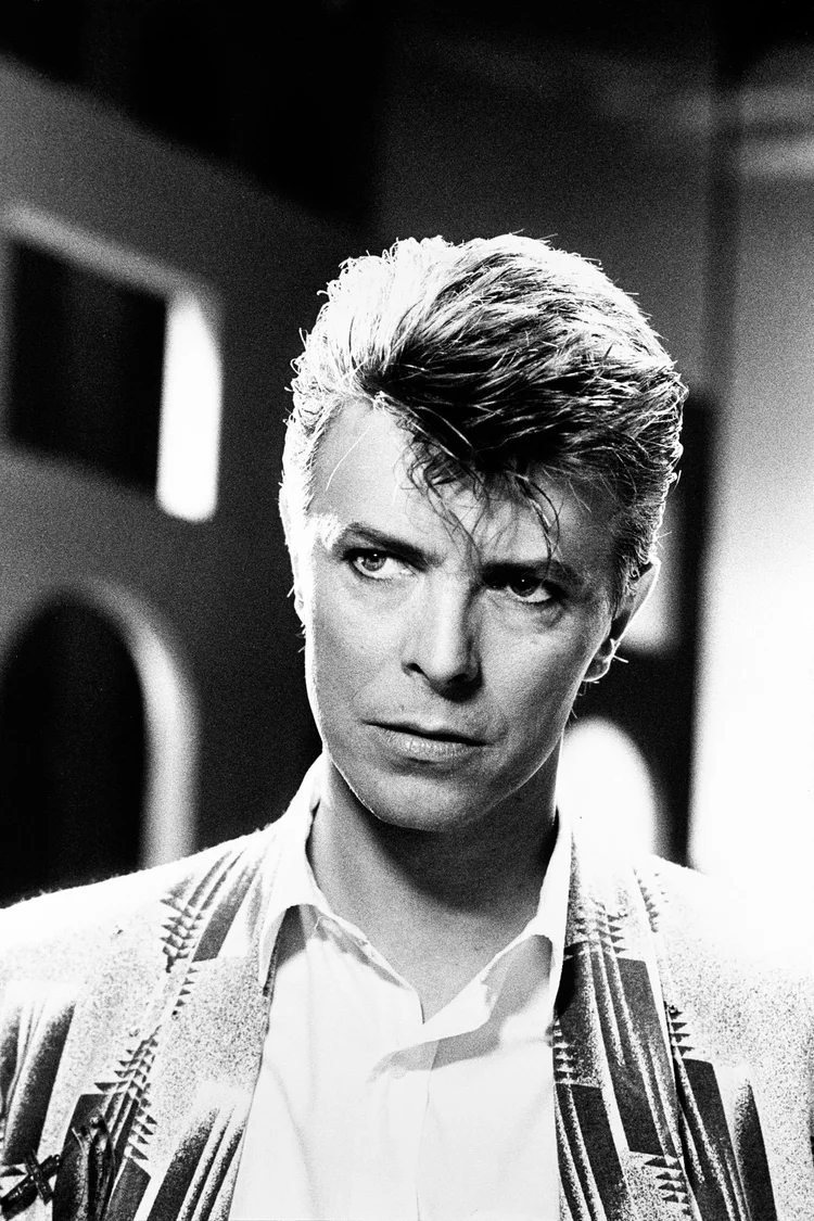 Black-and-white portrait of David Bowie looking intensely off camera, with styled hair, a white shirt, and a patterned jacket against a blurred urban background.