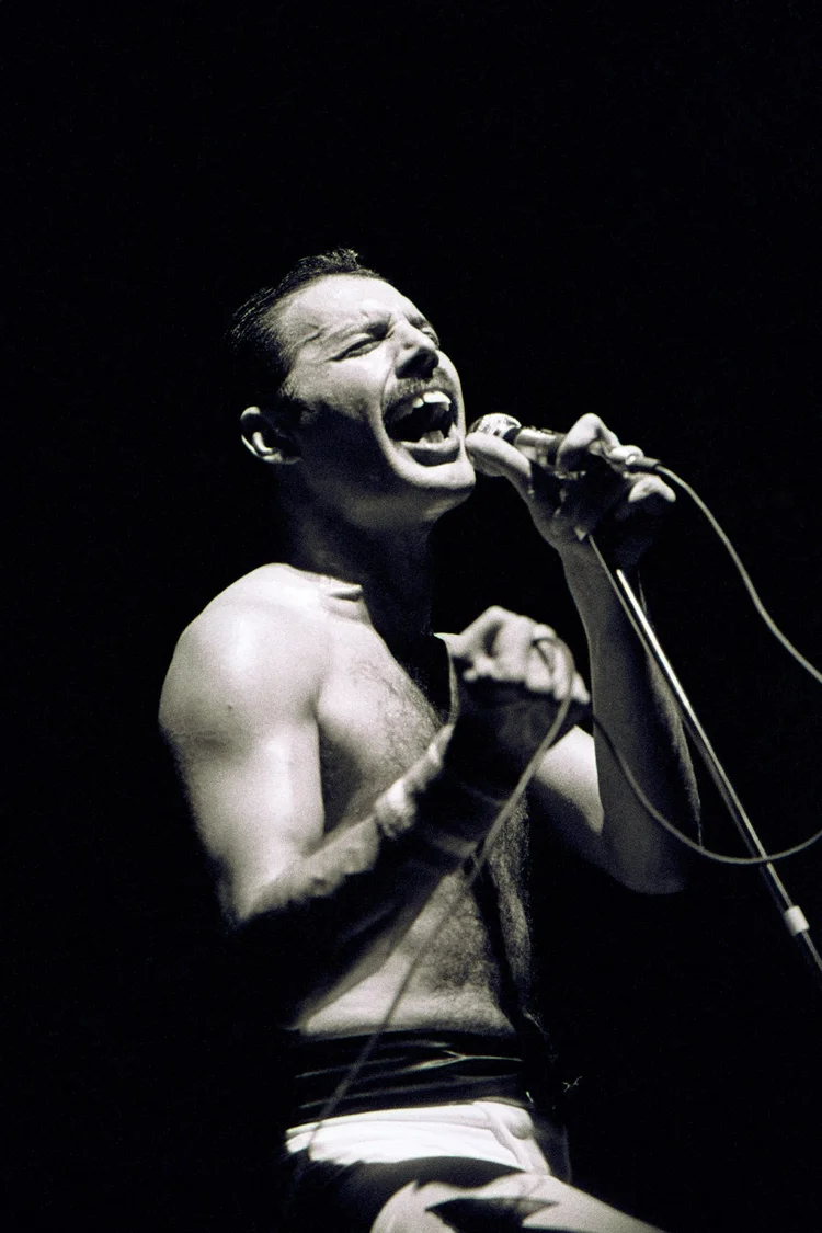 High-contrast concert photograph of Freddie Mercury singing into a microphone onstage, shirtless and mid-performance, against a dark background.