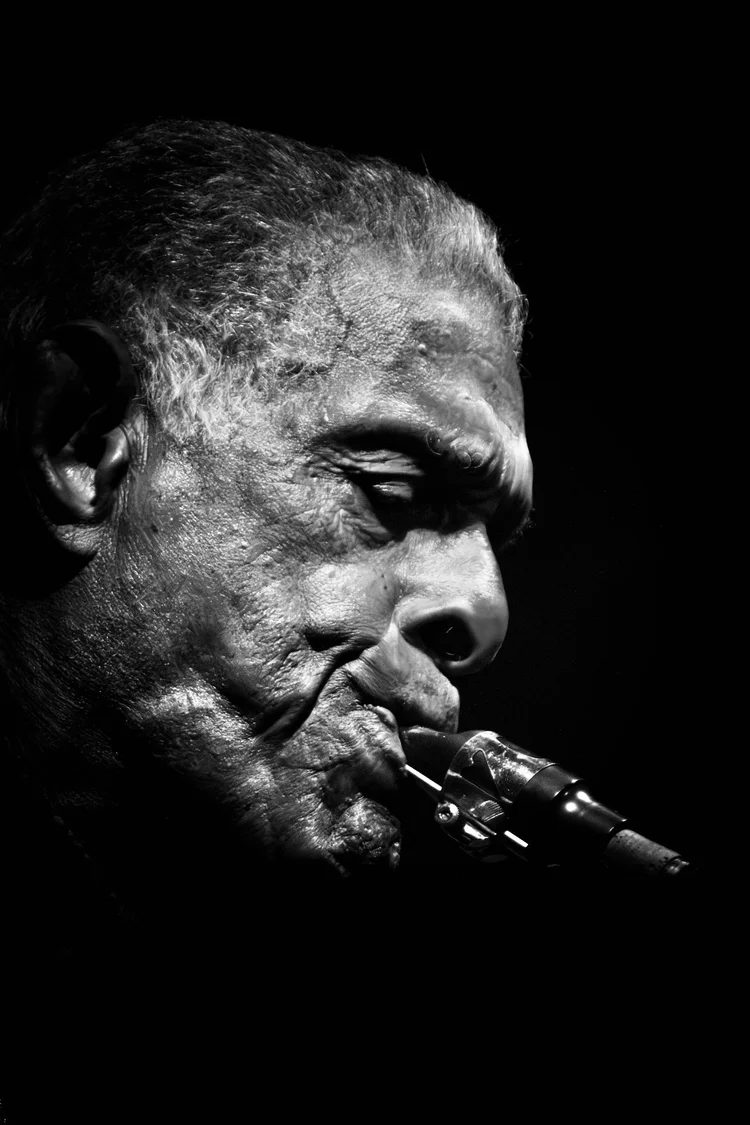 Black-and-white side-profile portrait of a Preservation Hall jazz musician playing a saxophone, with dramatic lighting highlighting the texture of his face against a black background.