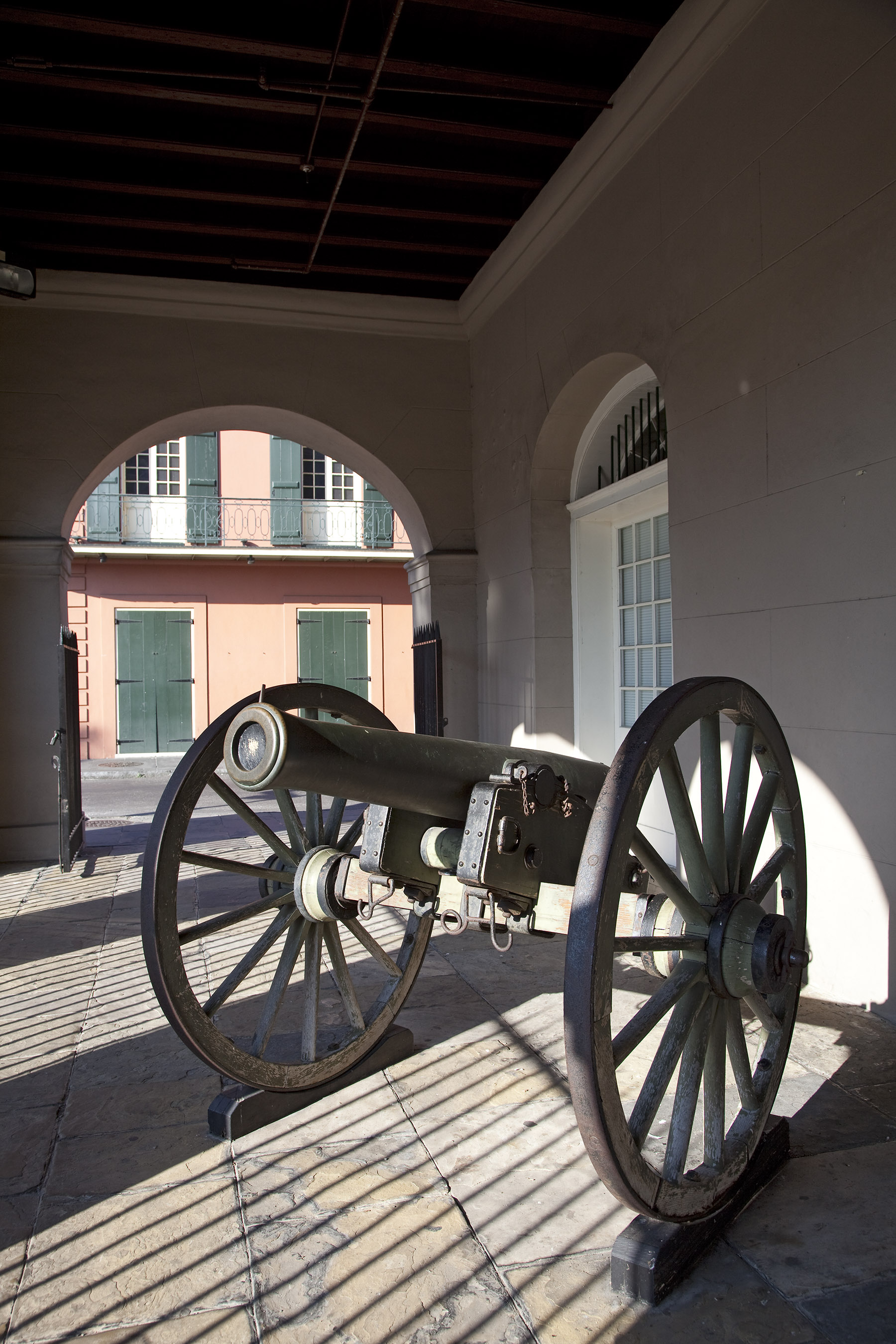 A historic cannon with large wooden-spoked wheels stands under the arched passageway of the Cabildo in New Orleans. Sunlight casts iron gate shadows across the stone floor, with a pastel building and green shutters visible through the archway.