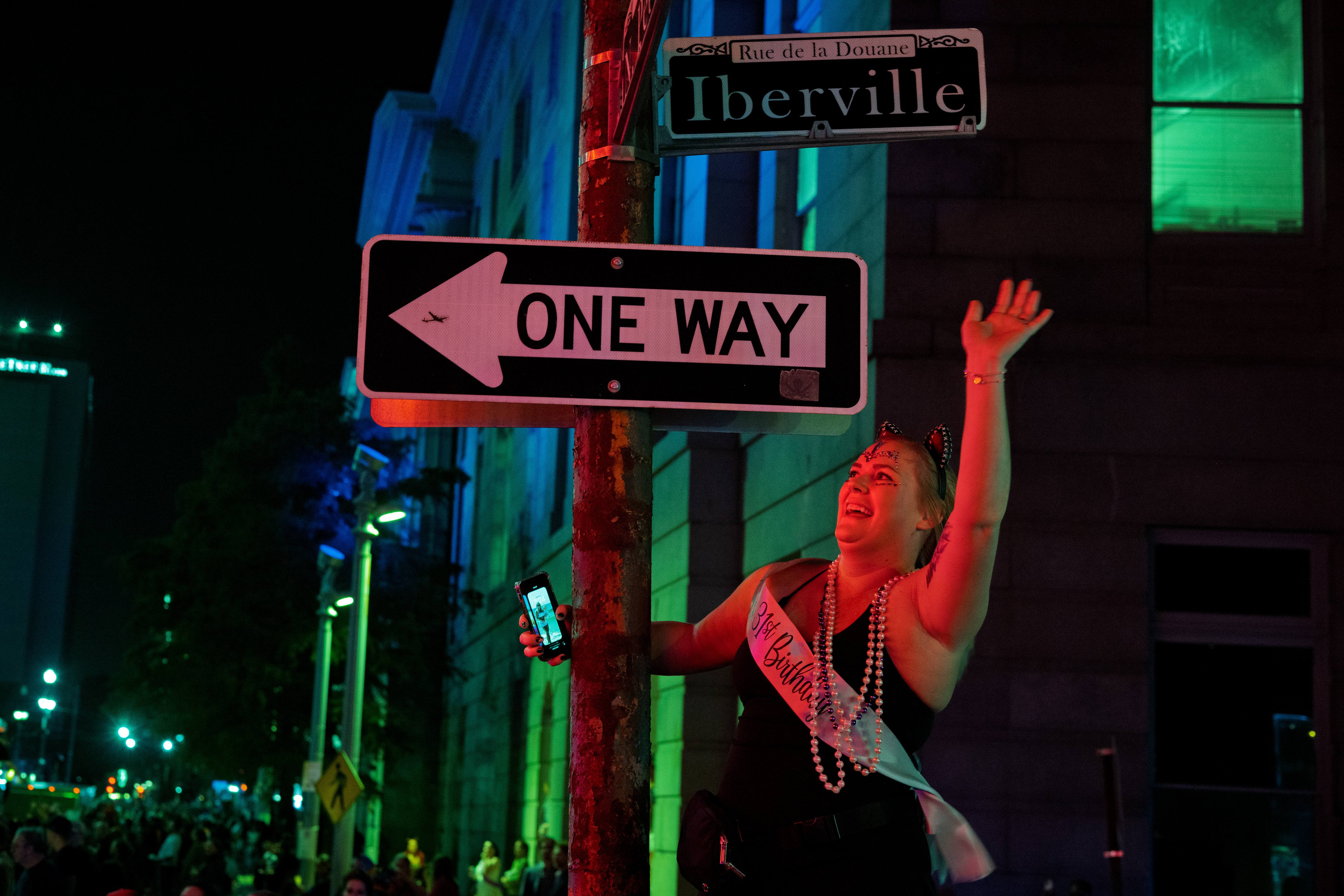 A woman wearing a birthday sash waves beneath a one-way sign at Iberville Street during a nighttime New Orleans street celebration, with colorful lights and a crowd in the background.