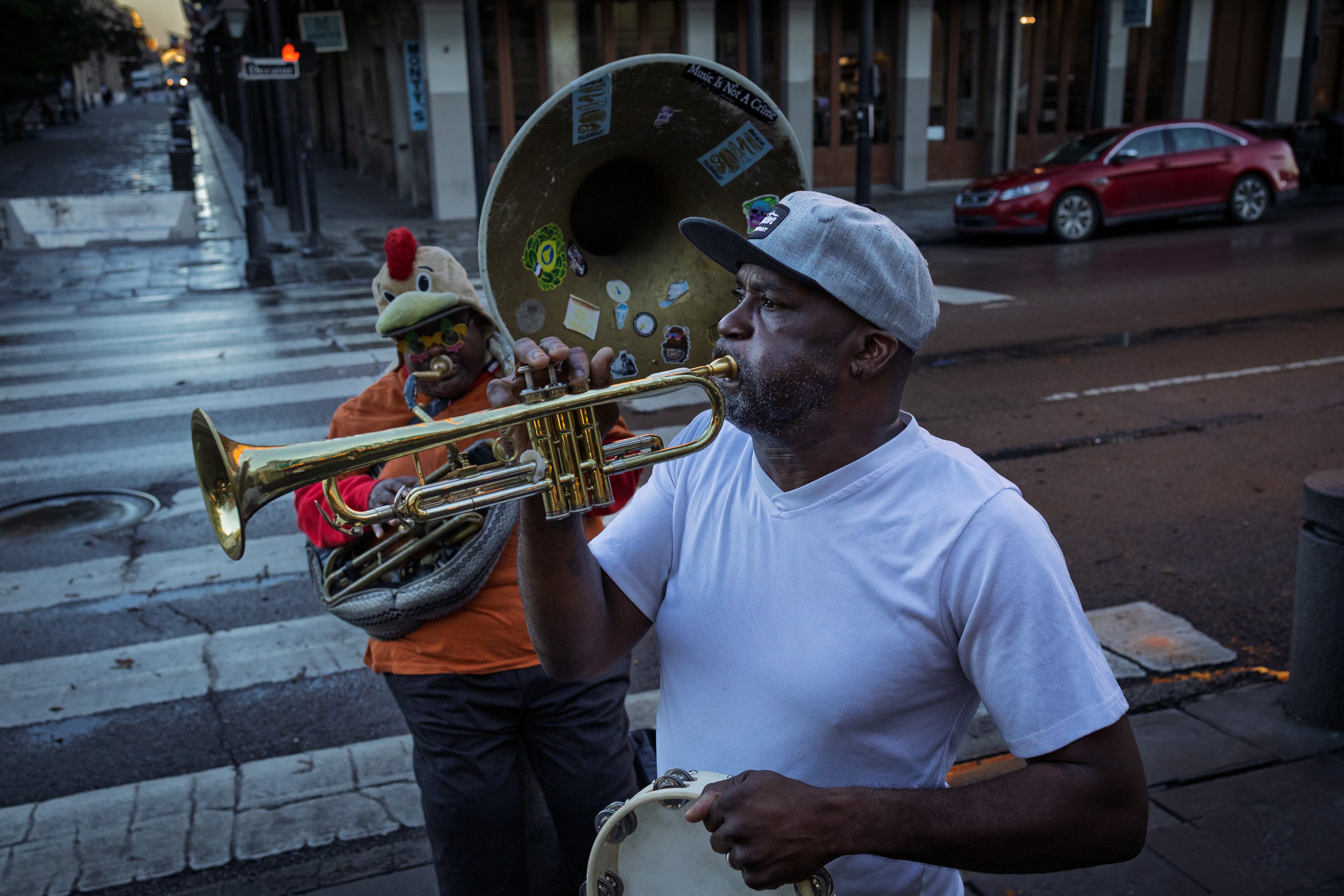 Two brass musicians perform in the street at dusk, one playing trumpet in the foreground and another playing sousaphone behind him.
