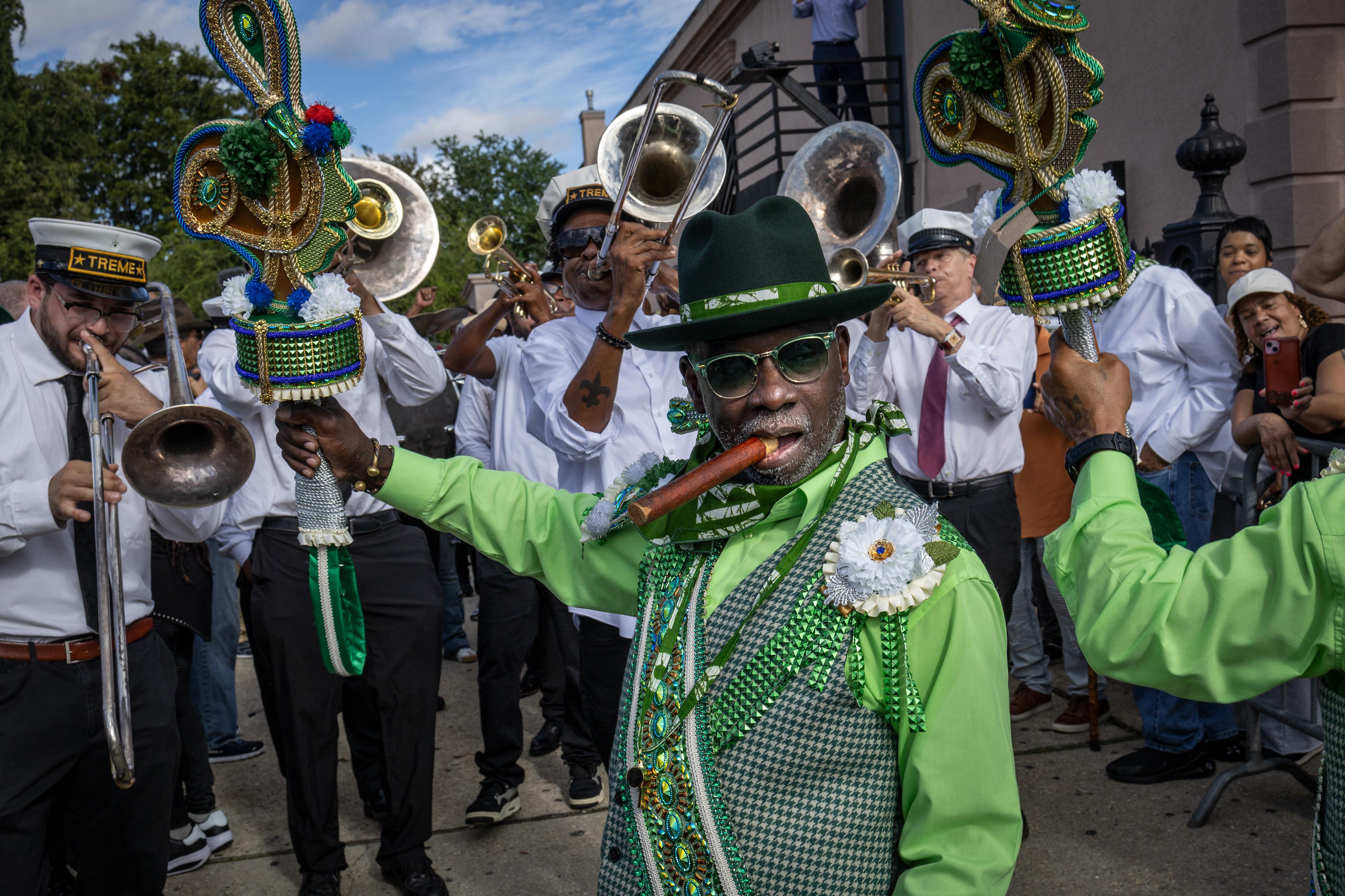 A man with a cigar in a bright green suit holds decorated fans while a brass band performs behind him during a New Orleans street procession.