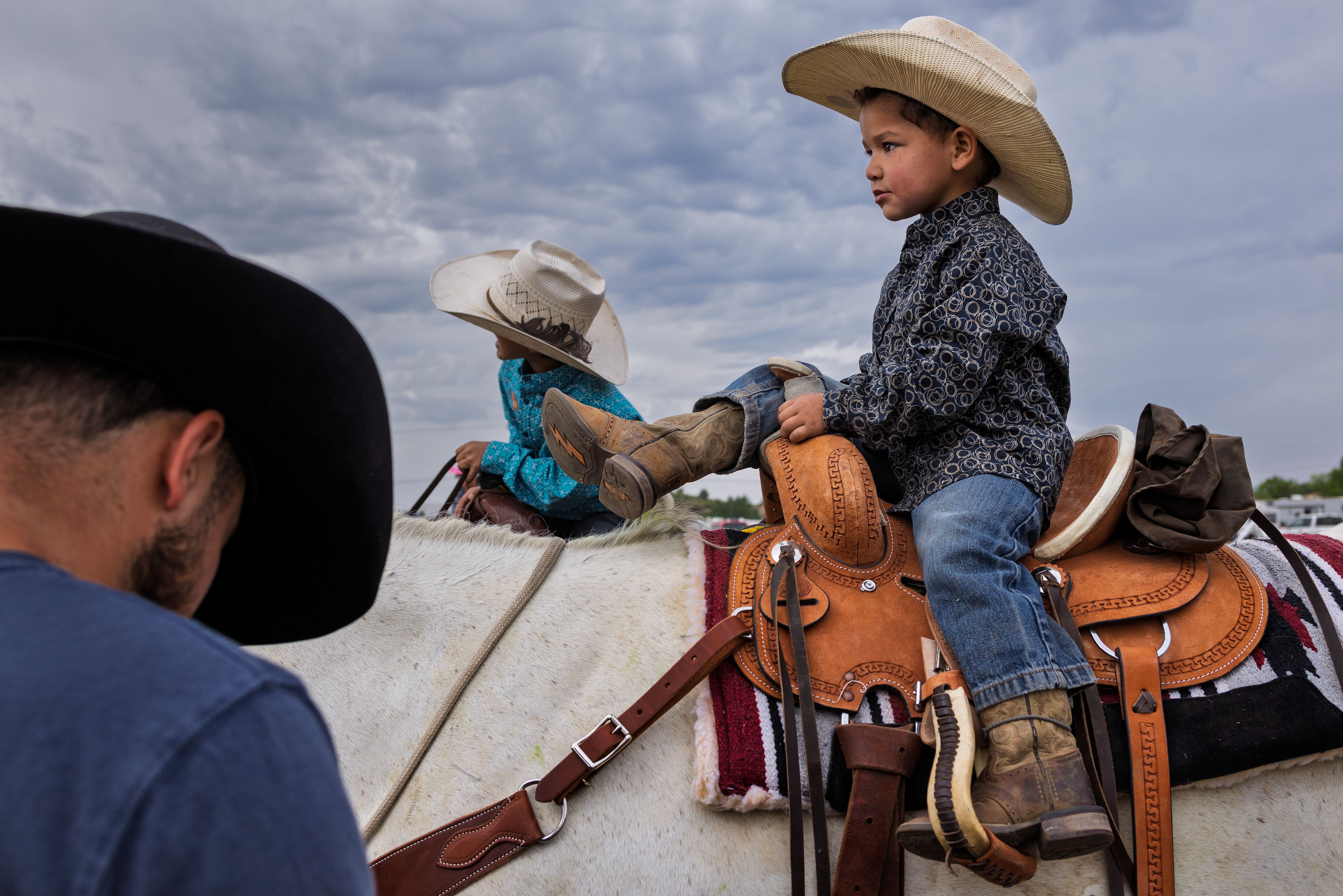 Two young children in cowboy hats sit on saddled horses under a cloudy sky, with an adult standing nearby.