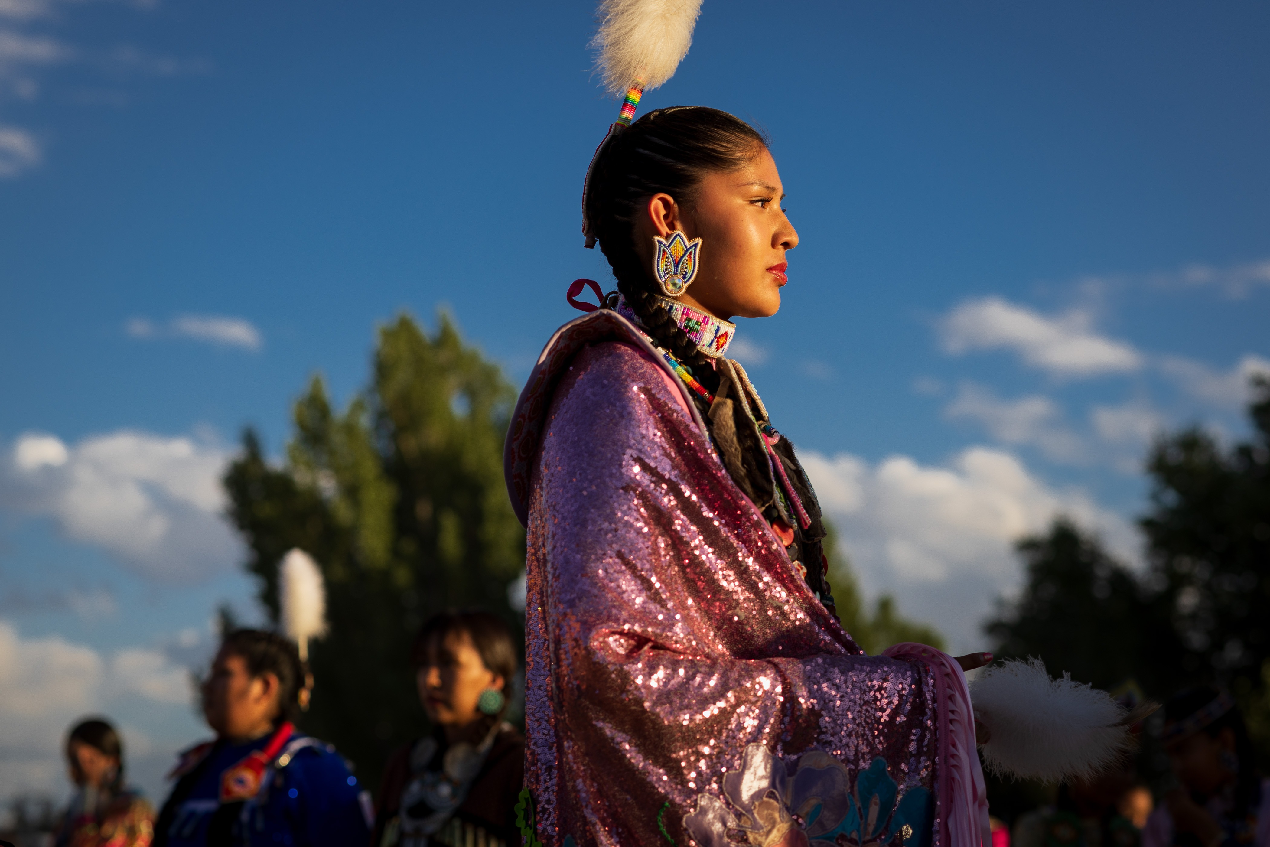A young Indigenous dancer in pink regalia looks into the distance during an outdoor gathering, with other dancers softly blurred in the background.