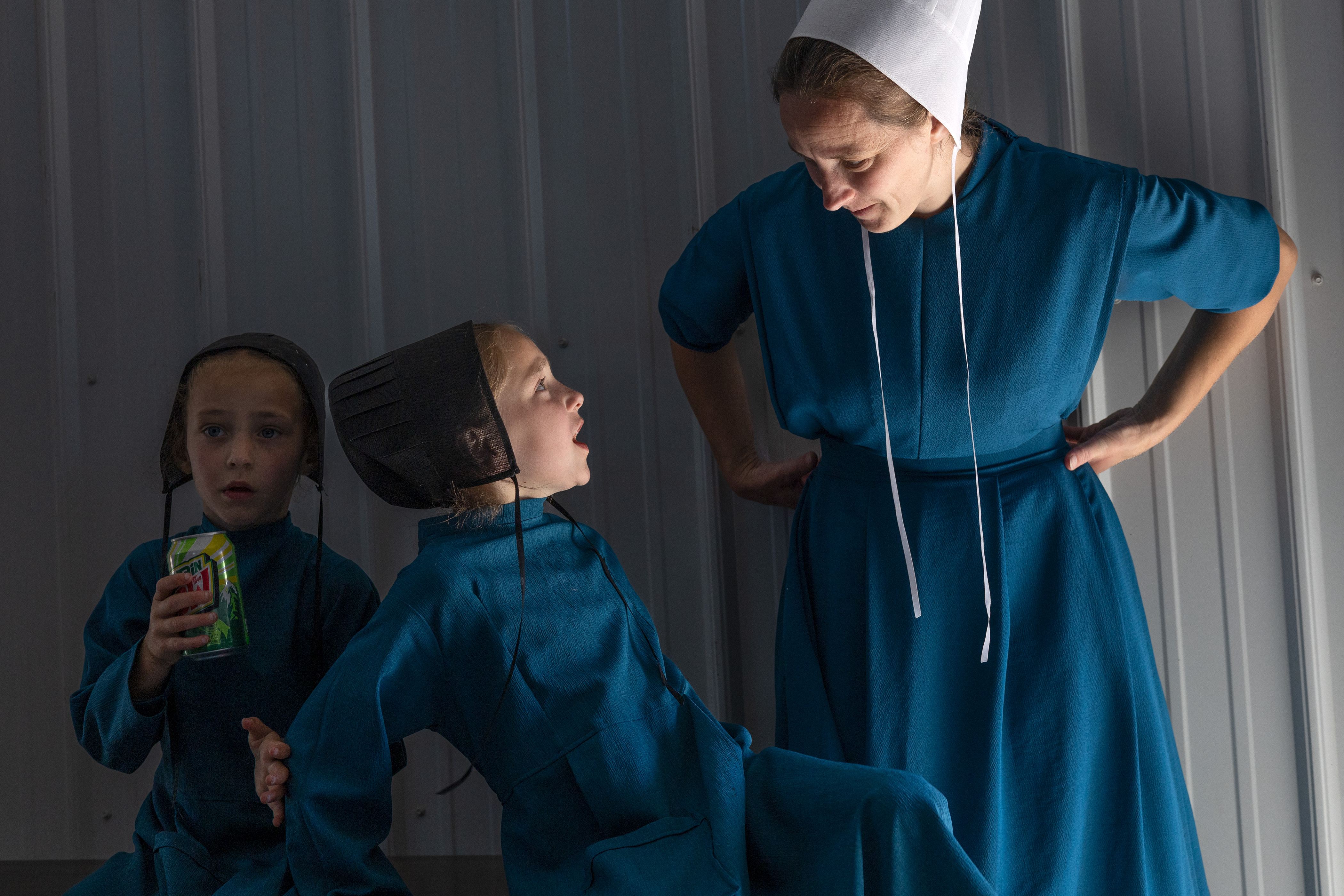 Two children in blue traditional clothing sit beside an adult in matching blue attire inside a simple room, one child looking up while speaking.