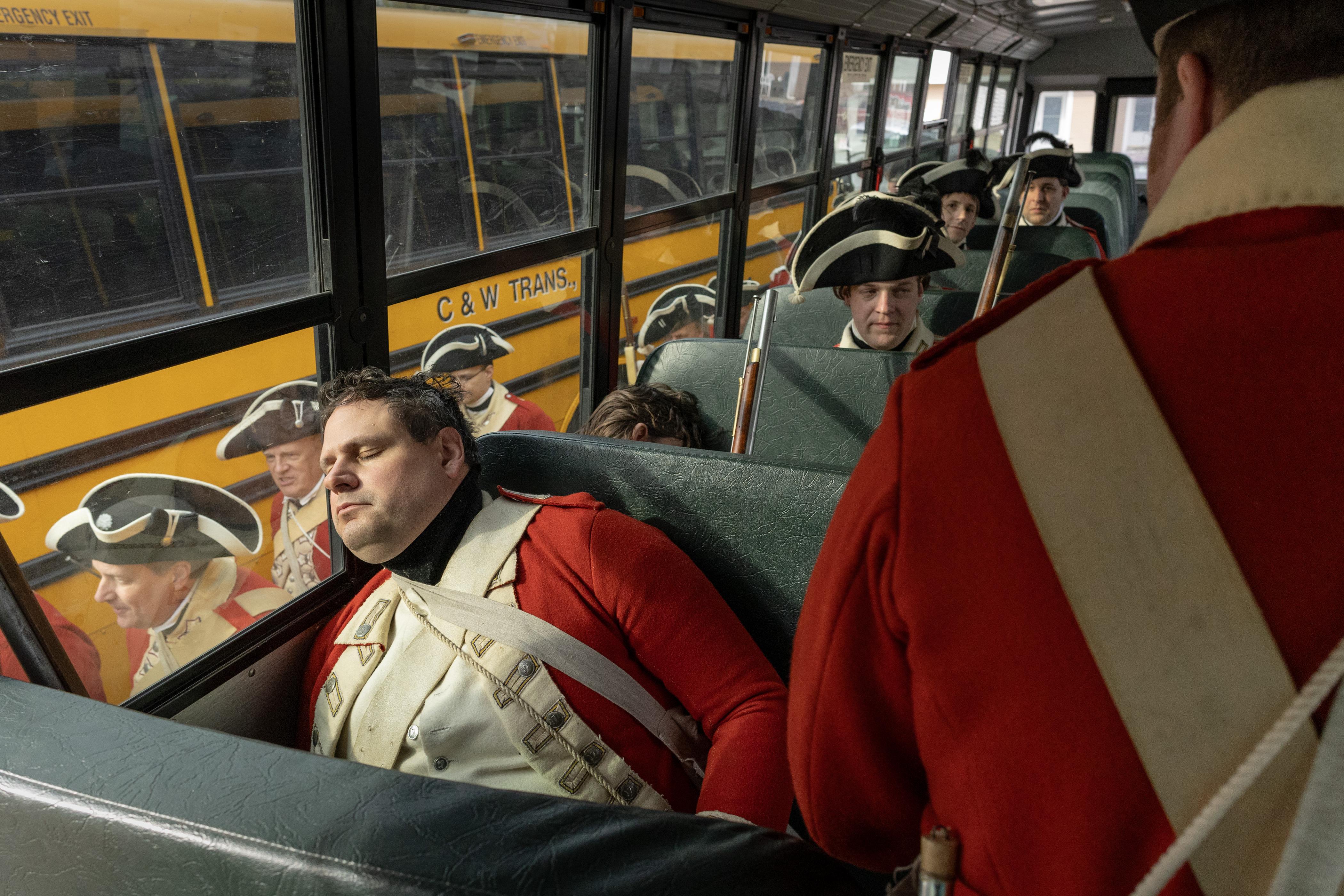 Historical reenactors dressed as British soldiers ride a bus, with one soldier asleep in the foreground and others seated behind him.