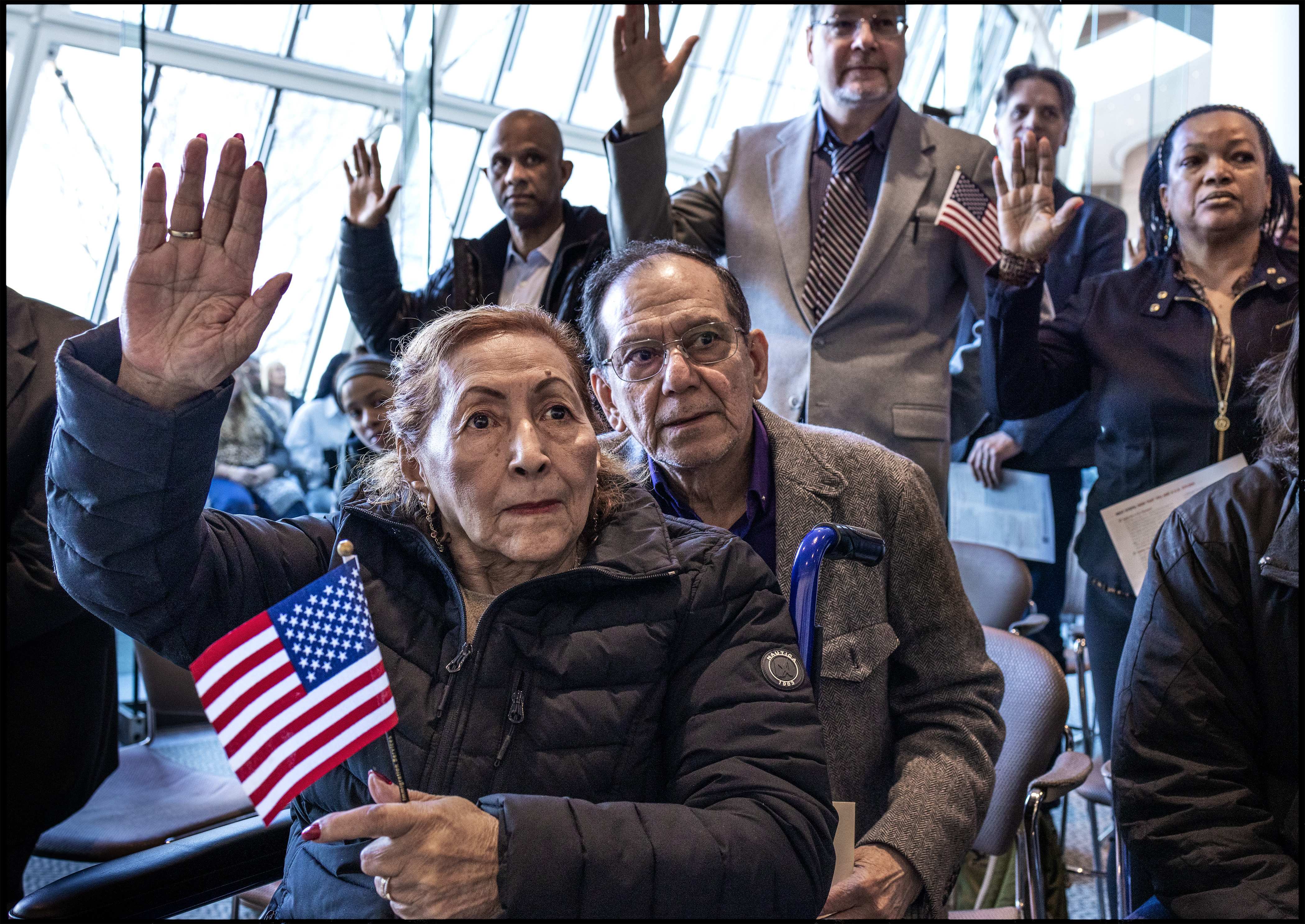 A group of people raise their right hands during a citizenship ceremony, with a woman in the foreground holding a small American flag.