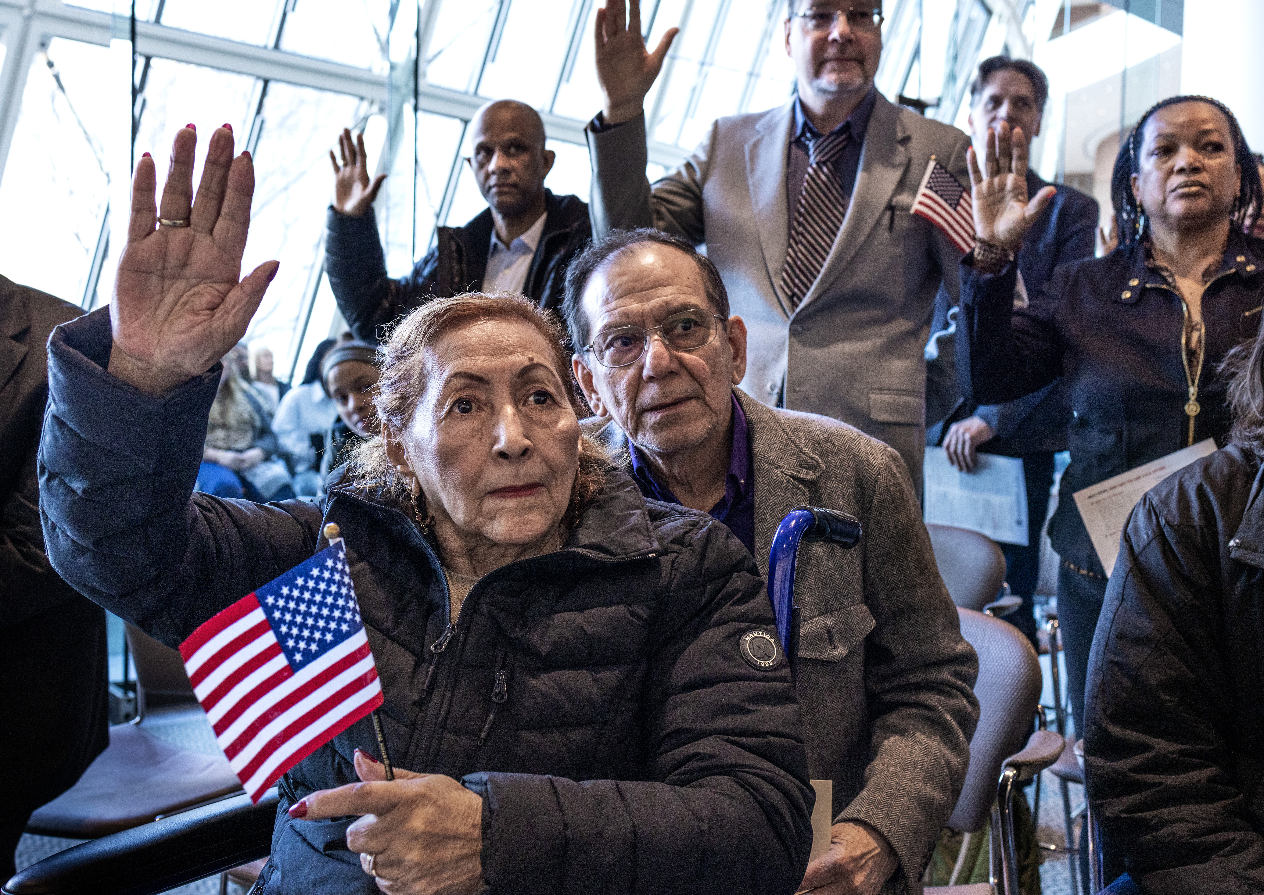 A group of people sit and stand in a bright glass-walled room, raising their right hands during a naturalization ceremony. In the foreground, an older woman holds a small American flag while raising her hand, with an older man seated beside her. Several other participants behind them also raise their hands, and one person has a small American flag tucked into his jacket pocket.