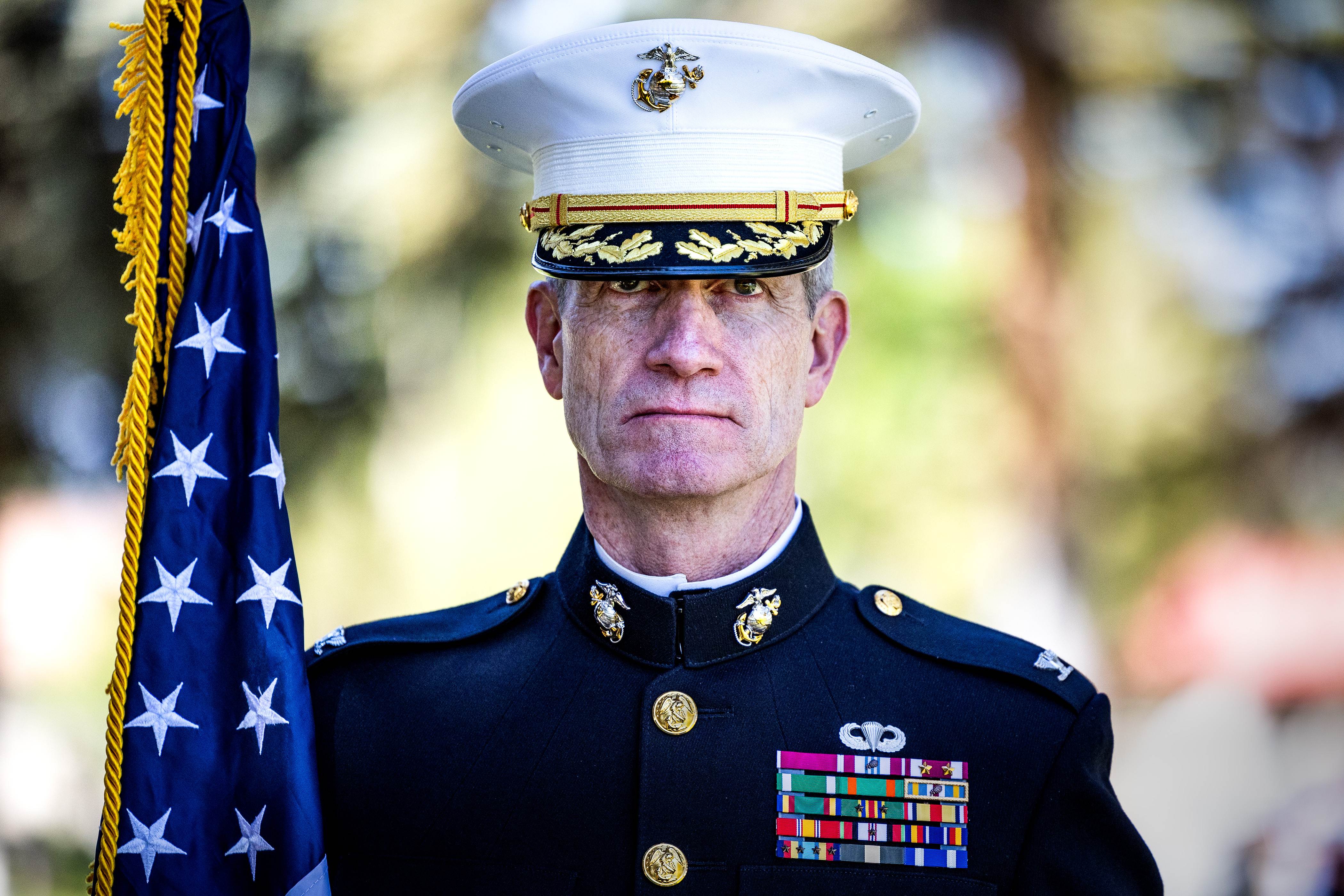 A U.S. Marine in dress uniform stands solemnly beside an American flag during an outdoor ceremony.