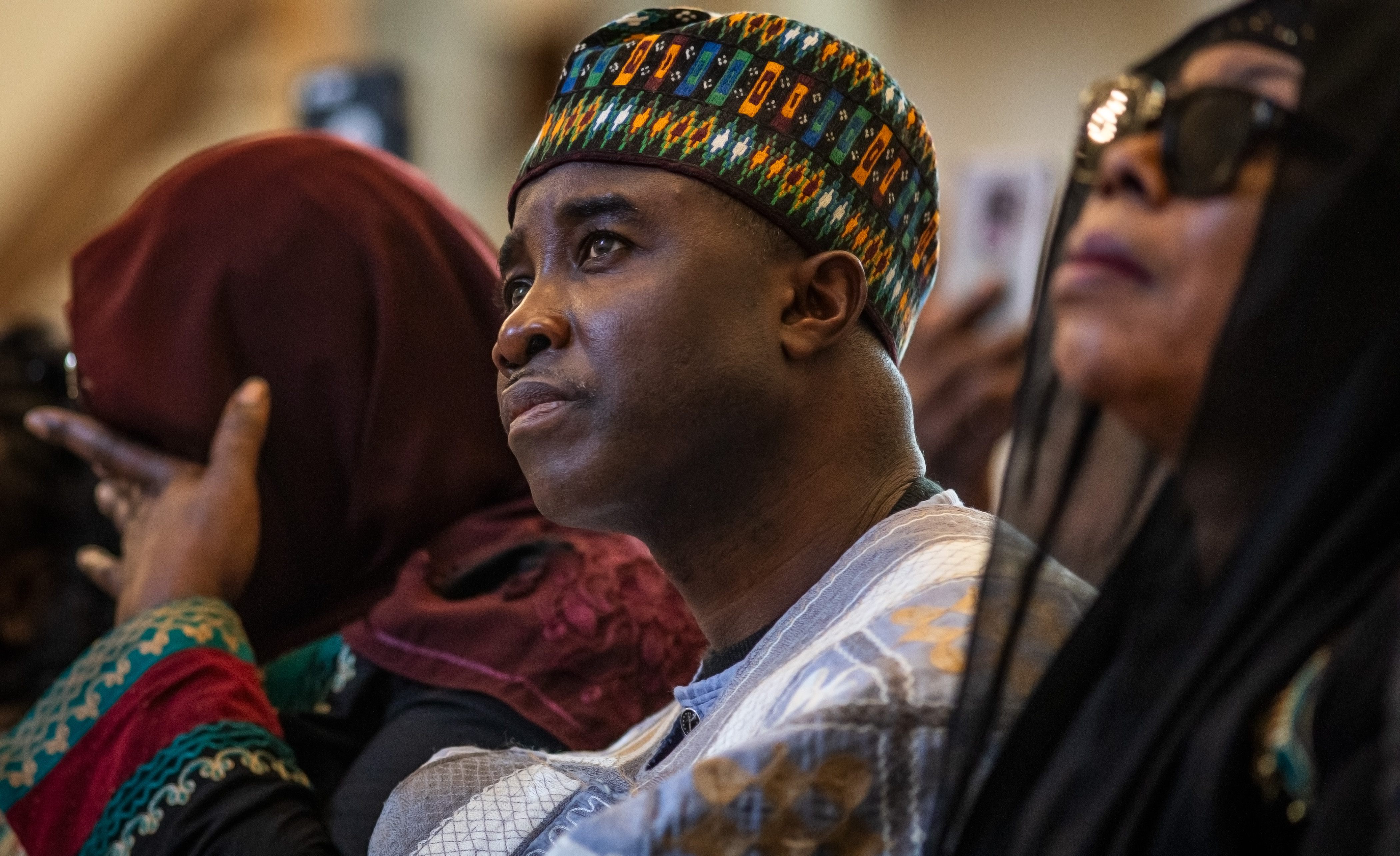 A man in a patterned cap and traditional clothing looks upward during an indoor ceremony, surrounded by other attendees.