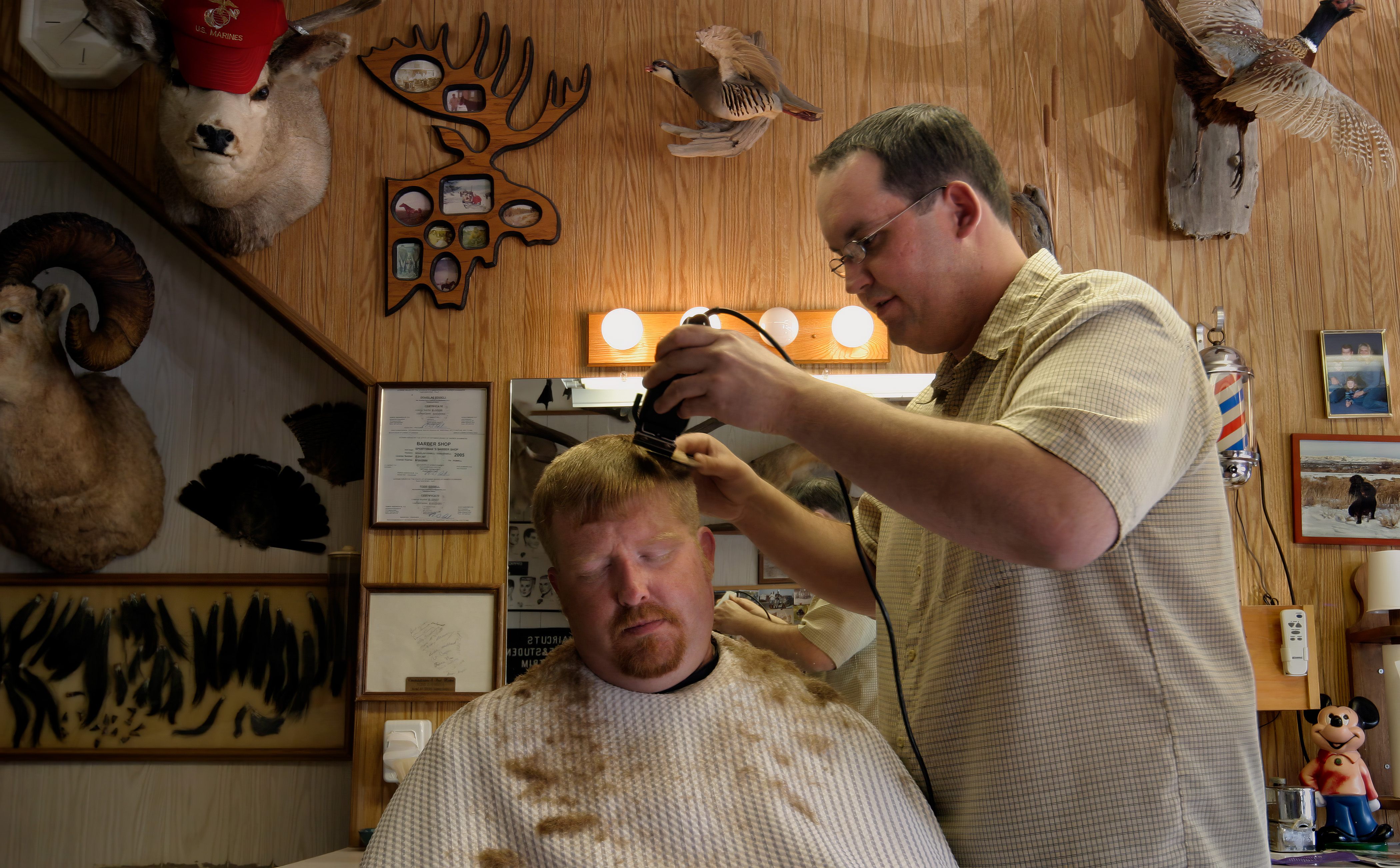 A barber cuts a man’s hair inside a wood-paneled barbershop decorated with taxidermy, framed photos, and memorabilia.
