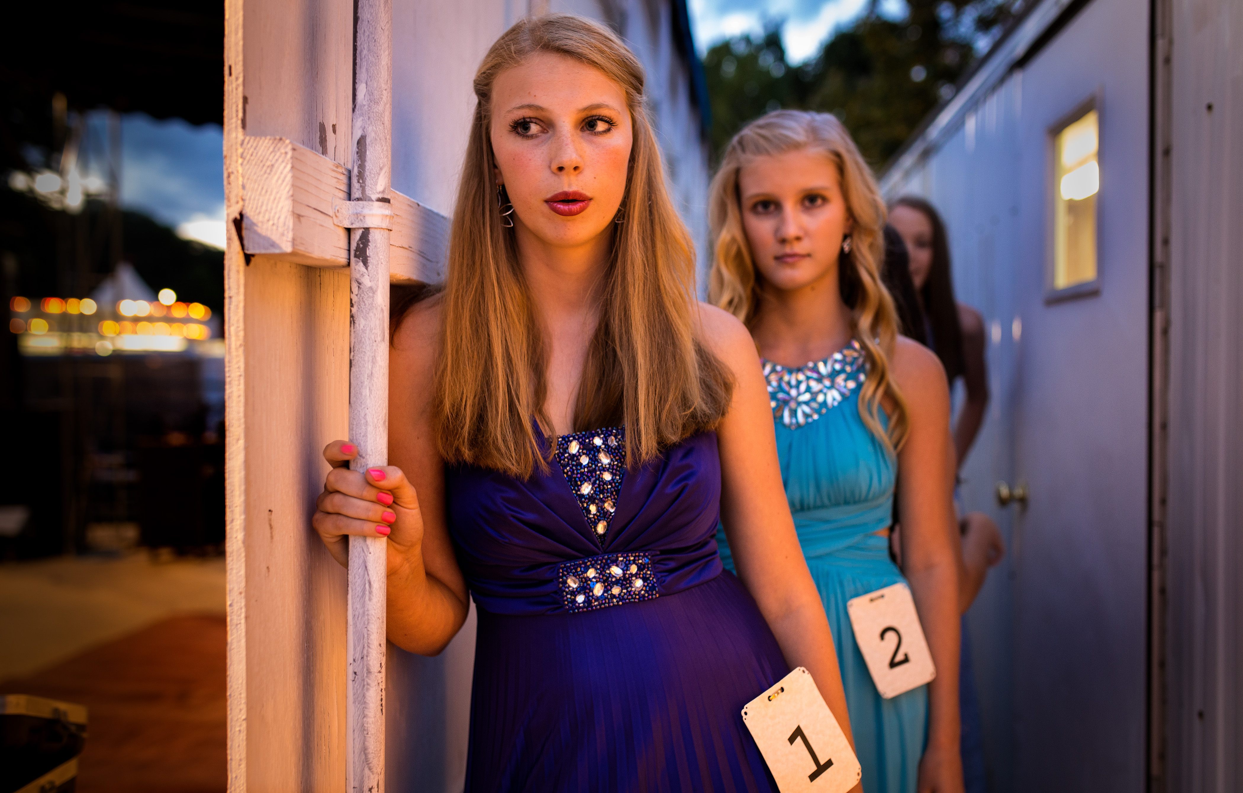 Young women in formal dresses wait backstage at an evening event, wearing numbered contestant tags.