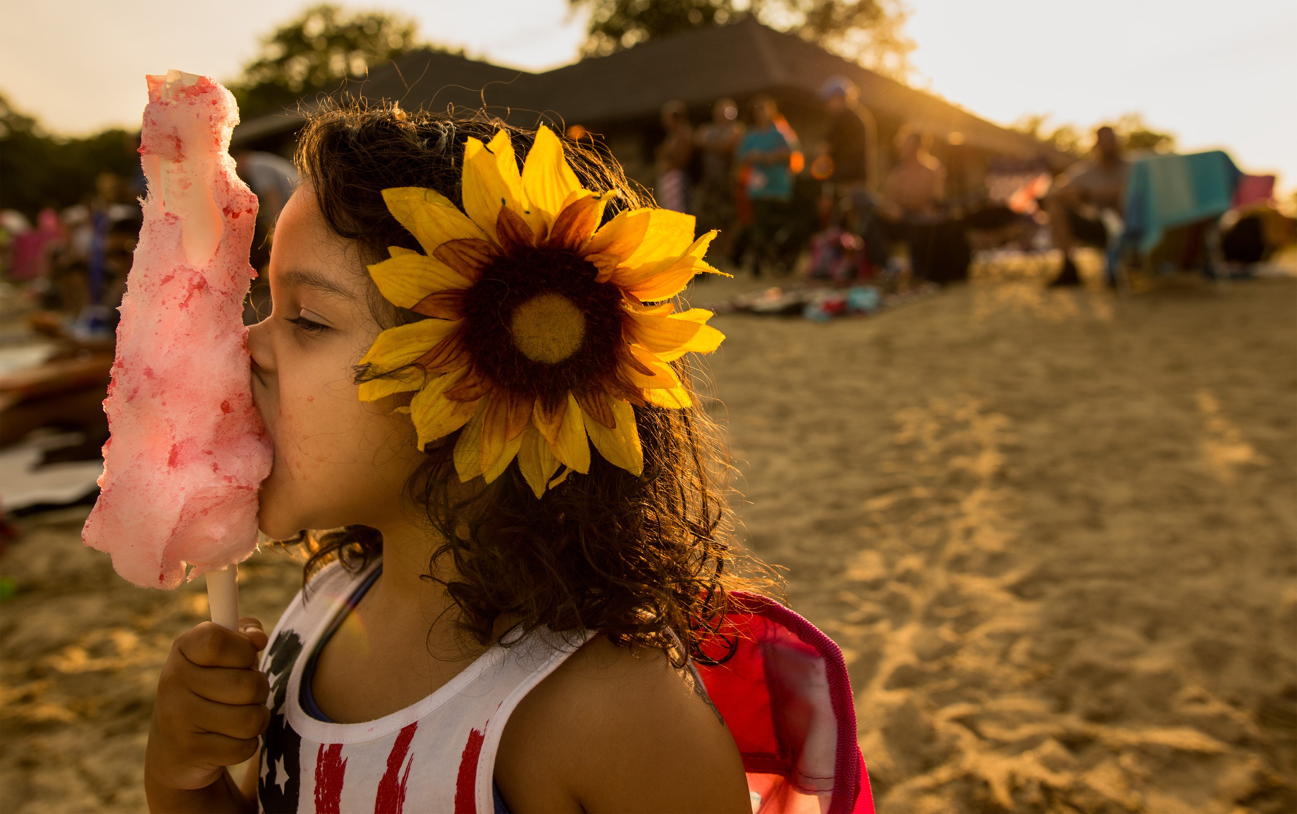 A child with a sunflower in her hair eats pink cotton candy at an outdoor fair or festival during golden-hour light.