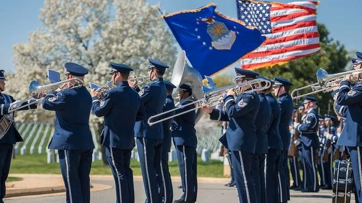 U.S. Air Force Band members in dress uniforms perform outdoors with brass instruments and drums. American and Air Force flags wave above the band, with rows of white headstones visible in the background.