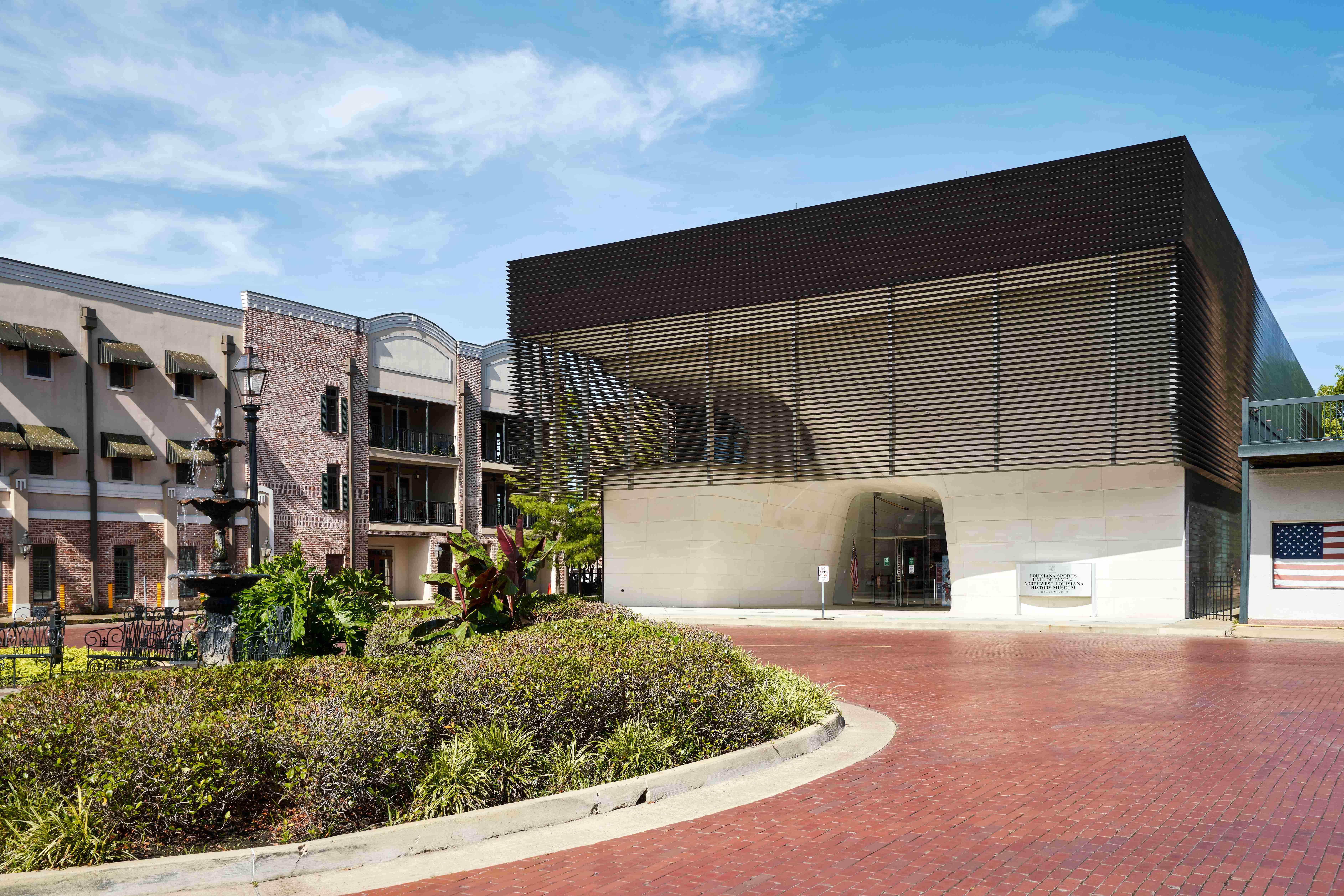 Exterior view of the Louisiana Sports Hall of Fame and Northwest Louisiana History Museum in Natchitoches, Louisiana. The modern museum building features a dark, louvered upper façade above a light stone entrance, set along a red brick street near landscaped planters, a decorative fountain, and neighboring brick buildings under a blue sky.