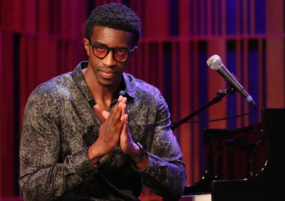 Kyle Roussel sits at a piano onstage beside a microphone, hands clasped, with warm red and purple lighting in the background.
