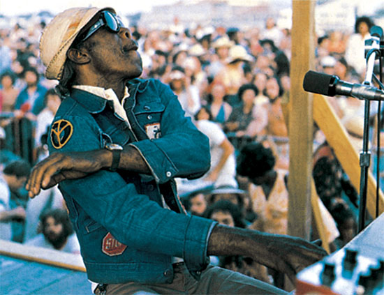 Professor Longhair performs at a piano on an outdoor stage, wearing sunglasses and a cap, with a large festival crowd in the background.
