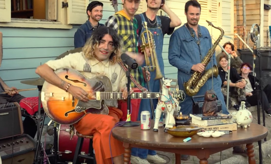 A band performs outdoors in front of a house, with a seated guitarist singing into a microphone while other musicians stand behind him holding brass and woodwind instruments.